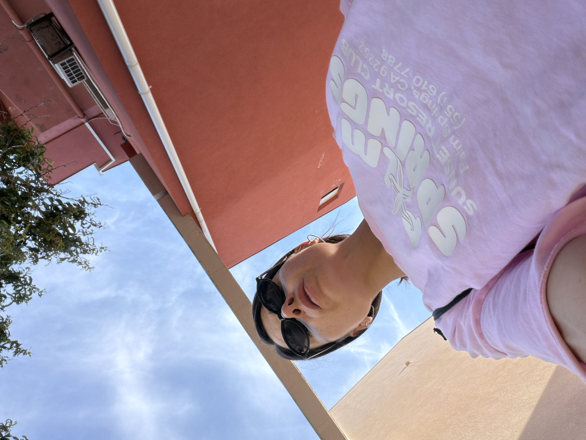 A low-angle outdoor shot shows a person wearing sunglasses and a pink shirt, standing outside near a building. The sky is blue with some white clouds. Parts of the building are salmon-colored, with visible ventilation structures, and tan. The person is wearing a pink shirt with the words "Sunset Resort Club, Palm Springs CA 92262, (551) 610-7788." The image does not include information about the location in Budva, Montenegro.