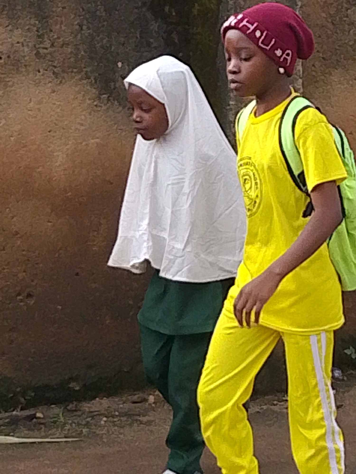 Two young girls are walking side-by-side on a dirt path with a textured, weathered wall in the background. The girl on the left is wearing a white hijab and a dark green skirt. She is looking downwards. The girl on the right is wearing a bright yellow t-shirt with a circular logo on it, bright yellow pants with white stripes down the sides, and a maroon beanie with white lettering that appears to spell "H U A". She is also wearing a light green backpack. Both girls appear to be walking away from the viewer, toward the right side of the frame. The environment suggests an outdoor setting, possibly in an urban or semi-urban area. Given the subject matter, this image was likely captured in Jos, Nigeria, a city known for its diverse population and religious communities where school uniforms and headscarves are common.