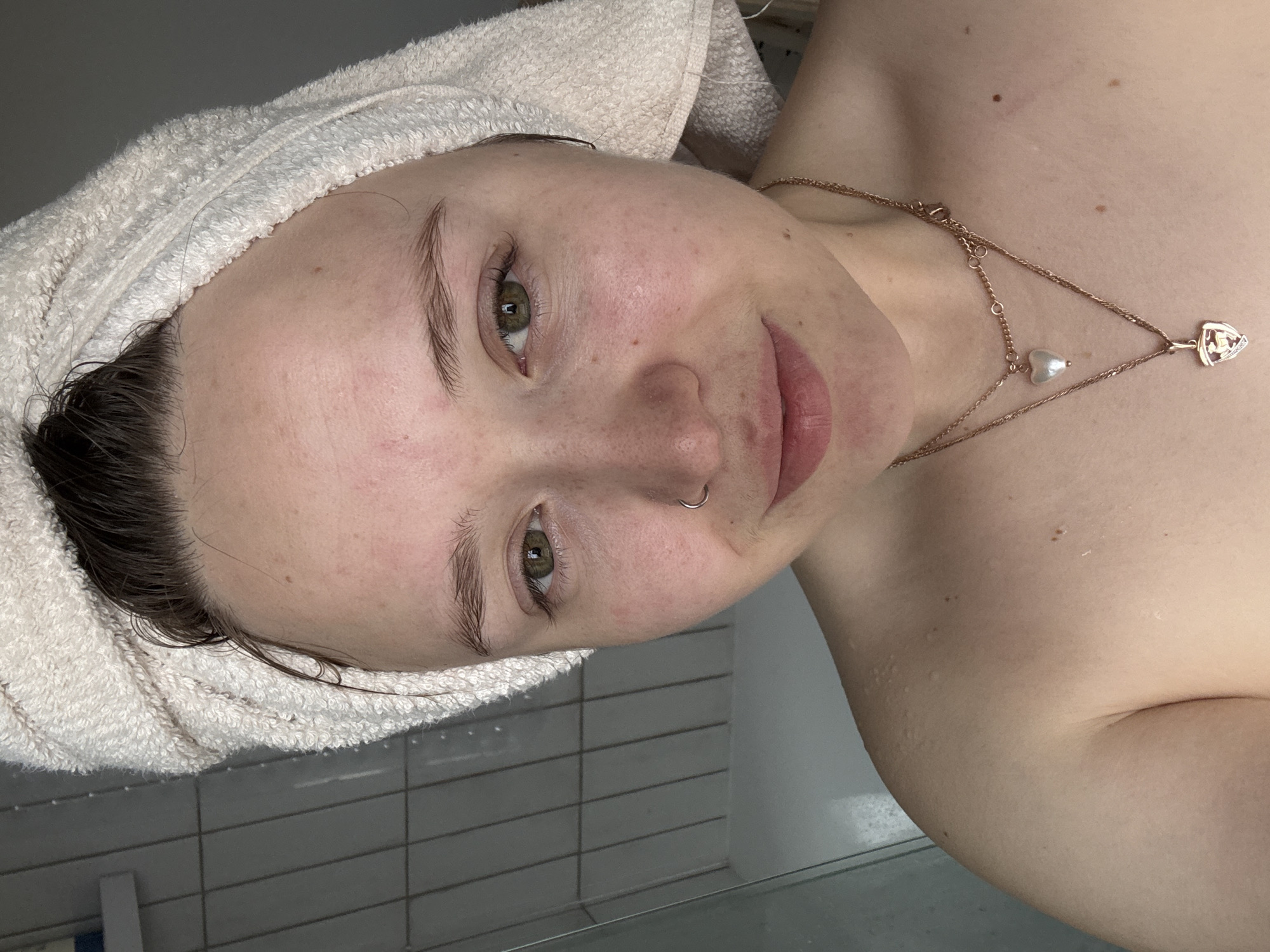 A medium close-up shot captures a person with fair skin and light features. A white towel is wrapped around the head, covering most of the hair. The face shows green eyes and the individual appears to have a nostril piercing. A delicate, gold-colored necklace adorns the neck, featuring a pearl and a heart-shaped pendant. The background includes a tiled wall and a hazy white surface, suggesting a bathroom or shower setting. There is no explicit indication of Izhevsk, Russia, in the image. The person seems to be looking directly at the camera with a slight smile.