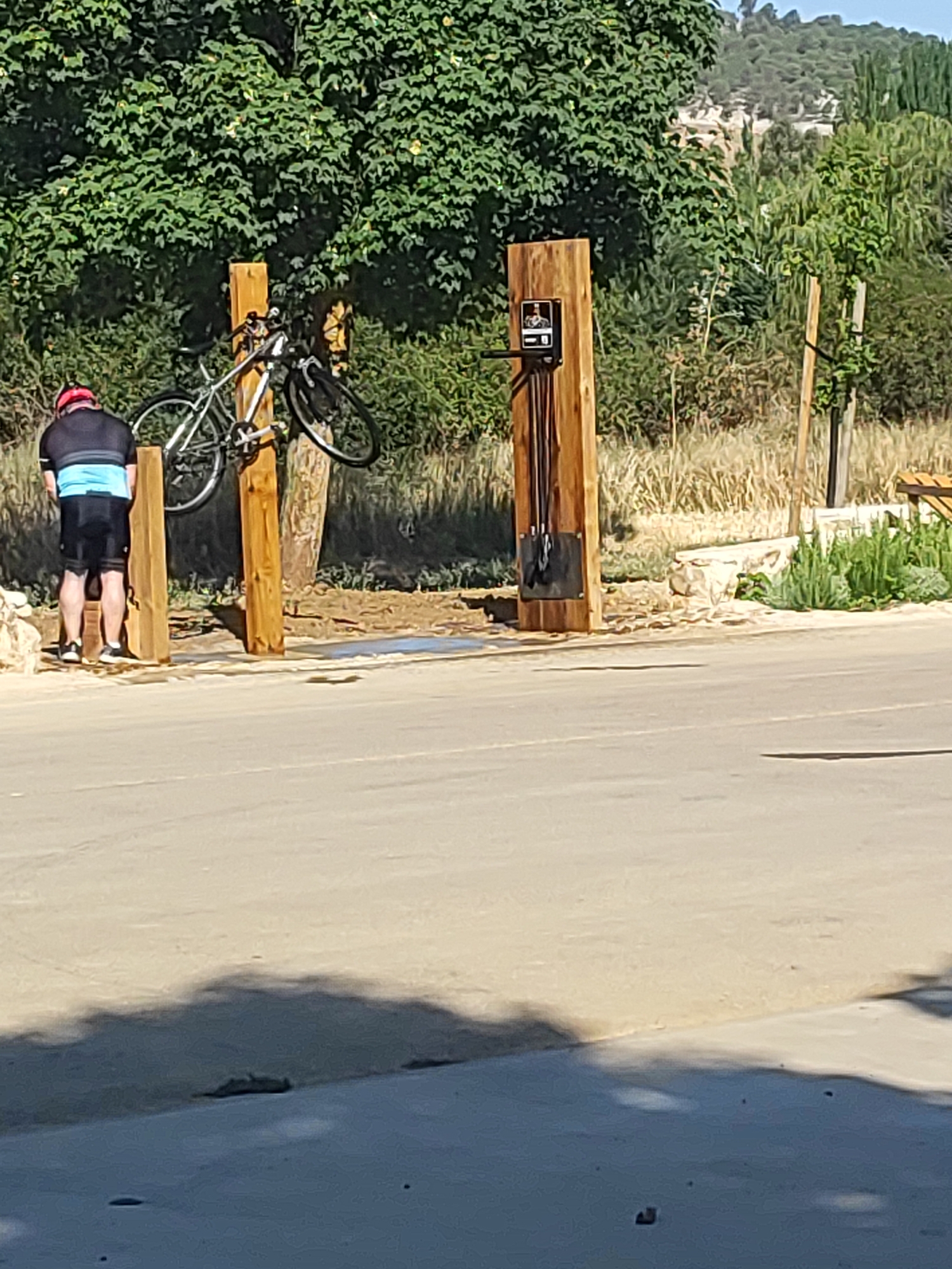 In Villamuriel de Cerrato, Spain, a cyclist in athletic attire stands next to a public bicycle repair station. The station, made of thick wooden posts, includes a mount for holding a bicycle upright and a panel with tools. A bicycle is suspended from one of the wooden posts. The cyclist, seen from behind, is wearing a red helmet, a dark jersey with a blue stripe, and black shorts. The environment is outdoors, with a paved surface in the foreground and a natural landscape with trees and vegetation in the background. The lighting suggests it is daytime.