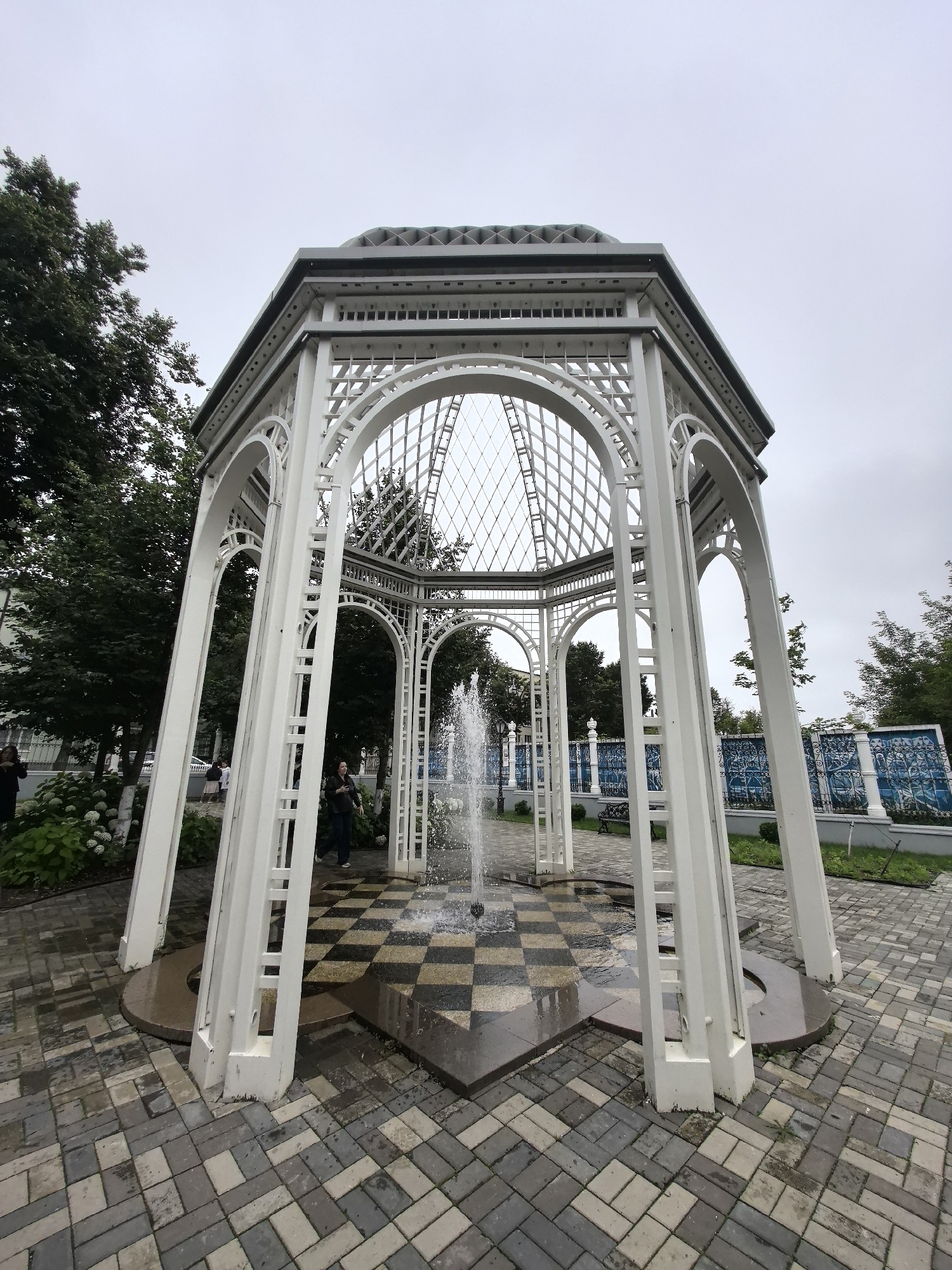 This is a wide-angle, eye-level, outdoor photograph of a white octagonal gazebo with an ornate lattice roof. A fountain is active in the center of the gazebo, with water spraying upwards. The ground inside and immediately around the gazebo is paved with alternating dark and light square tiles, creating a checkerboard pattern in some areas. The area surrounding the gazebo is paved with interlocking rectangular pavers in shades of gray and beige. To the left of the gazebo, there are lush green trees and a white hydrangea bush with white blooms. In the background, there is a blue ornamental fence with a repetitive pattern. Several individuals are visible; one person is standing within the gazebo, looking to their left, and another person is walking past the gazebo on the left side. The sky is overcast and gray. This gazebo is located in the city of Perm, Russia, specifically in the Perm Botanical Garden.