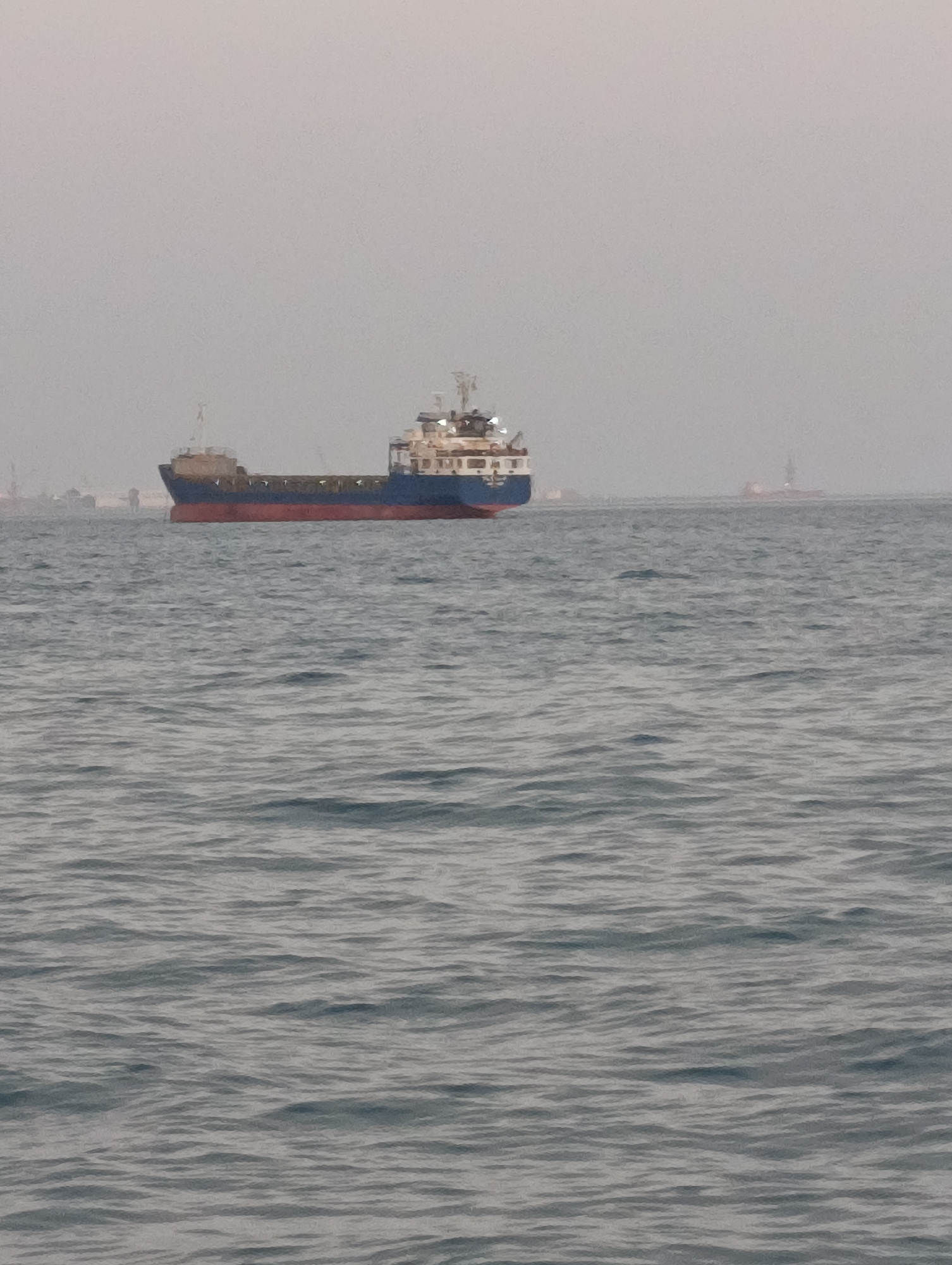 A cargo ship is anchored in the Atlantic Ocean, off the coast of Setúbal, Portugal. The ship, which appears to be a bulk carrier, is painted blue and red, with a white superstructure. It is laden with cargo, as indicated by the cranes and other equipment visible on deck. The sea is calm, with a light chop. The sky is overcast, with a thick layer of clouds obscuring the sun. In the distance, a few other ships can be seen, as well as the silhouette of the city of Setúbal. The image is shot from a low angle, with the ship taking up the majority of the frame. The overall impression is one of peacefulness and tranquility.