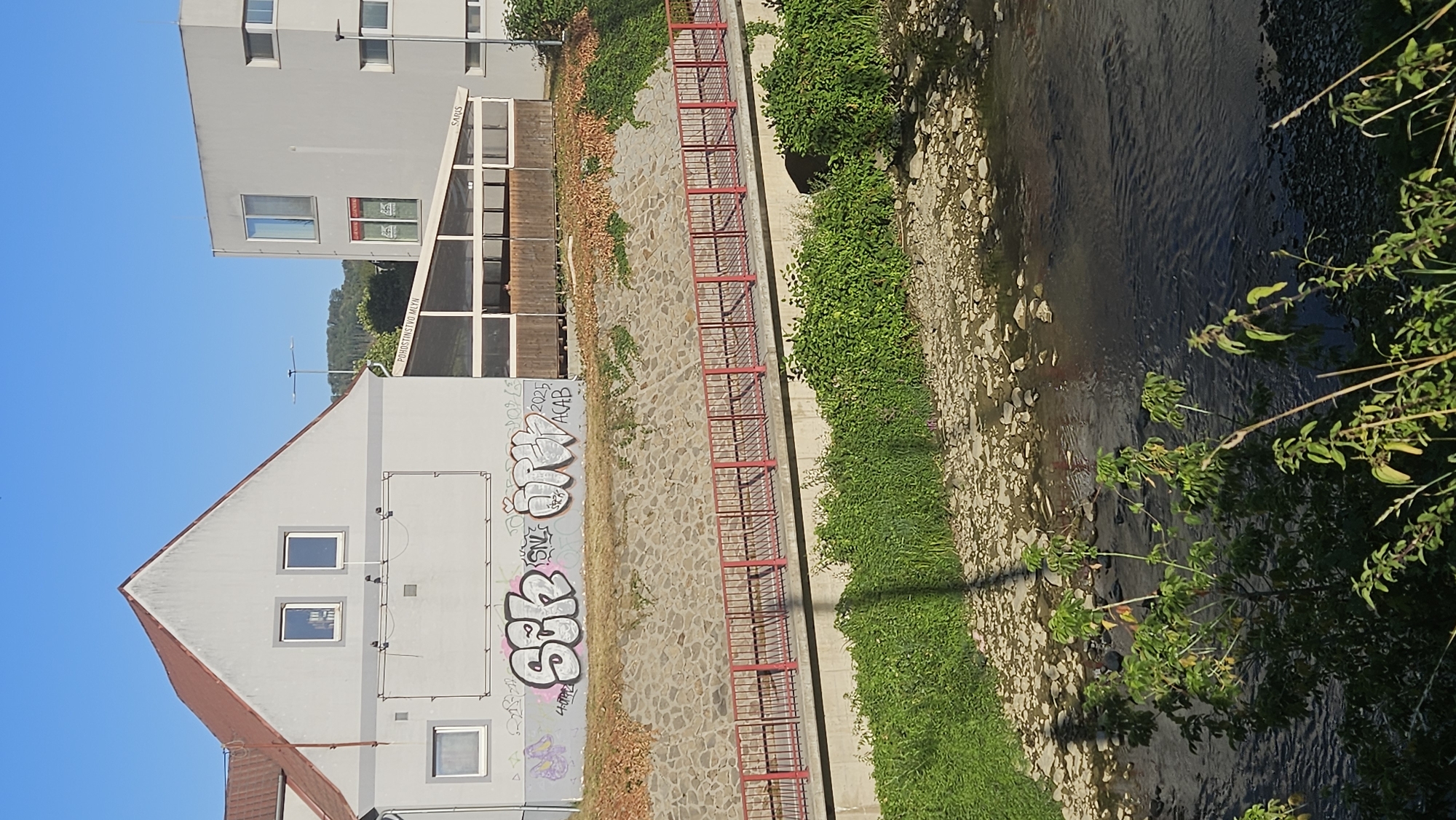 The image depicts an outdoor scene in the city of Prešov, Slovakia, likely along the Torysa River. In the foreground, a shallow riverbed with numerous small stones is visible, with dark water flowing over them. Lush green vegetation grows along the riverbank. A concrete embankment is topped by a walkway with a red metal railing. Behind the walkway is a stone retaining wall partially covered in ivy.
On the embankment, there is a light-colored building with a prominent gable roof and a red tile covering. Two square windows are set high on the gable, and a large rectangular panel, possibly a former advertising space, is situated below them. The building's lower section features graffiti, including bubble-style lettering in white and gray, and some pink spray paint. A smaller square window is visible to the left.
Adjacent to this building, a modern, multi-story, light gray building stands to the right. Between the two buildings, a covered outdoor seating area with a wooden fence and glass panels is visible. A sign reading "POHOSTINSTVO MLÝN" is affixed to the roof of this covered area, along with another sign that appears to say "SARIS." The sky above is clear and blue, with the sun casting shadows across the scene. The overall impression is of an urban environment integrated with a natural water feature.