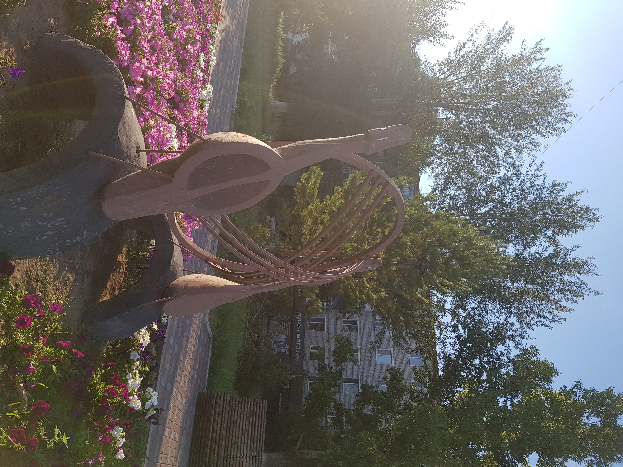 The image depicts an outdoor scene with a sculpture in the foreground, surrounded by flowers. The sculpture appears to be made of metal and has a reddish-brown patina, resembling a musical instrument with a circular body and a neck. It is placed near a flower bed filled with vibrant pink, purple, and white flowers.
Behind the sculpture, a paved pathway or sidewalk is visible, with a wooden fence or structure to the right. Further in the background, a brick building with multiple windows can be seen, partially obscured by lush green trees. The building has text written vertically on its facade, which reads "ДУЦЕН МАГАЗИН" (Dutsen Magazin).
The time of day appears to be late morning or early afternoon, as indicated by the bright sunlight casting lens flares across the image and the shadows. The sky is clear and blue, suggesting fair weather. The presence of blooming flowers and green foliage indicates a warm season, likely spring or summer. The scene is set in Kurchatov, Kazakhstan, as indicated by the location context. There are no people visible in the image, and the overall mood is serene and peaceful, highlighting the juxtaposition of nature and man-made elements.