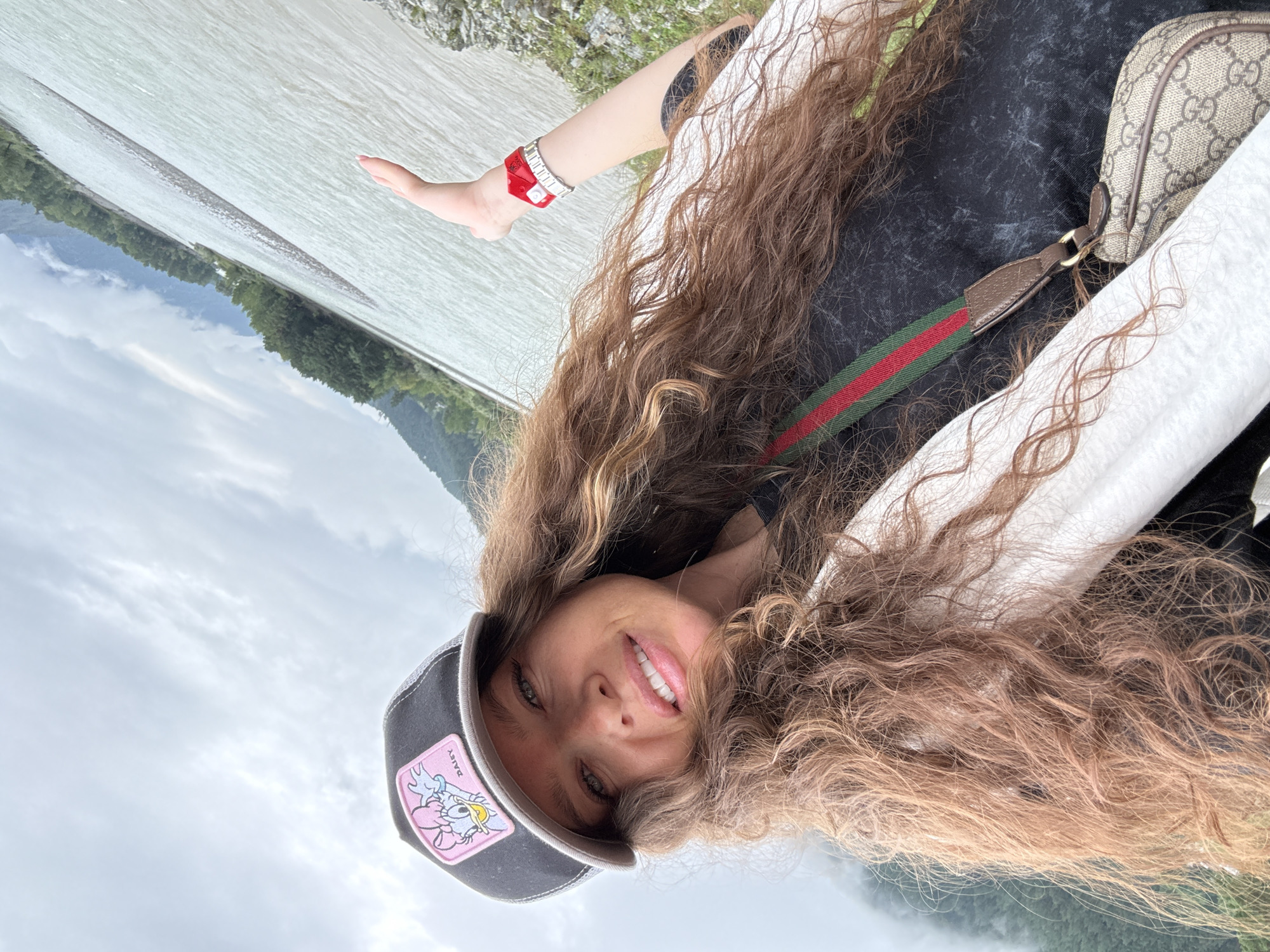 In Elekmonar, Russia, a young woman with long, wavy brown hair smiles at the camera while wearing a black baseball cap with a pink patch featuring a cartoon mouse. She is also wearing a white jacket over a black shirt, with a Gucci crossbody bag. Her left hand is extended, displaying a red bracelet. The background shows a wide, shallow river with a pebble bank, surrounded by green trees and mountains under a cloudy sky.