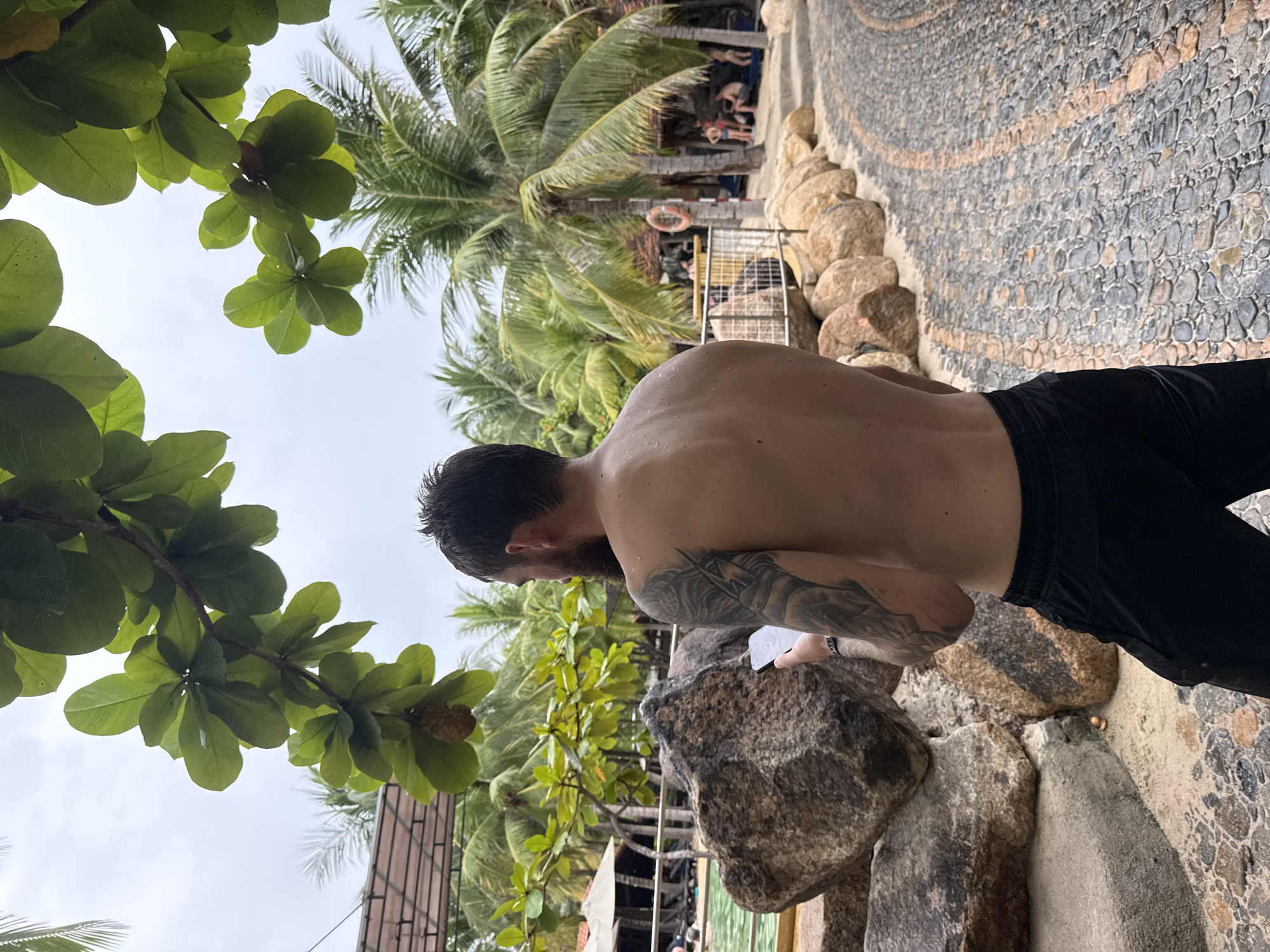 The image depicts a bare-chested man with tattoos on his left arm, wearing black athletic shorts. He is standing outdoors, holding a white object, possibly a phone or paper, in his right hand. He is facing away from the camera, looking towards a resort-like environment characterized by palm trees, a swimming pool with lounge chairs, and a paved walkway made of stones. The foliage overhead suggests a tropical setting. The presence of palm trees and the overall ambiance are consistent with a resort in Nha Trang, Vietnam, known for its beaches and tropical climate. The man appears to be alone in the immediate foreground, while other people are visible in the background near the pool area. The lighting suggests an overcast day.
