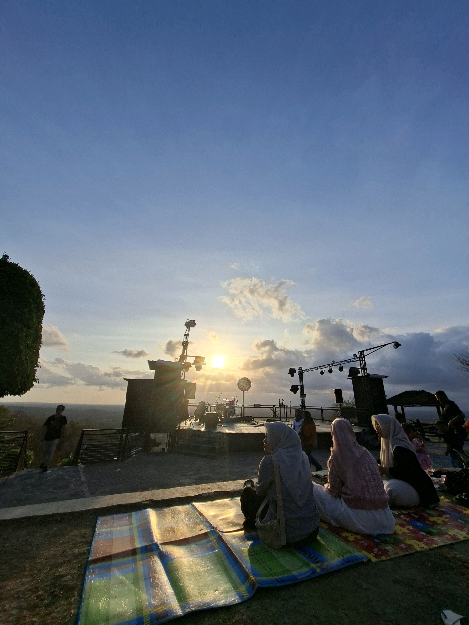 In Pleret, Indonesia, a group of people are gathered at an outdoor venue during sunset. The sun, low on the horizon, casts a warm glow and creates lens flare across the scene. In the foreground, several individuals, including women wearing hijabs, sit on colorful mats on the ground. In the mid-ground, a stage setup is visible with lighting equipment and what appears to be a drum set. A lone figure stands on a walkway in the left mid-ground, looking out at the expansive, hazy landscape below. The background features a vast sky with scattered clouds, typical of a tropical sunset. The overall atmosphere suggests a relaxed gathering or possibly an event taking place at dusk.
