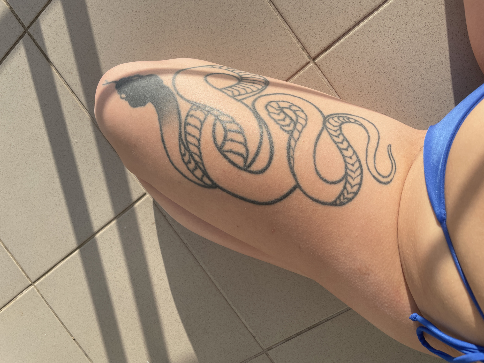 The image shows a person's leg with a snake tattoo, positioned on a tiled surface. The tattoo covers a significant portion of the thigh, depicting a stylized snake with a dark, filled-in head. A blue bikini bottom is partially visible on the right side of the image. The leg is positioned against a light-colored tile floor with shadows cast by parallel lines, possibly from an overhead structure. The location is indicated as Rafailovići, Montenegro, though there are no features that confirm the location within the image itself.