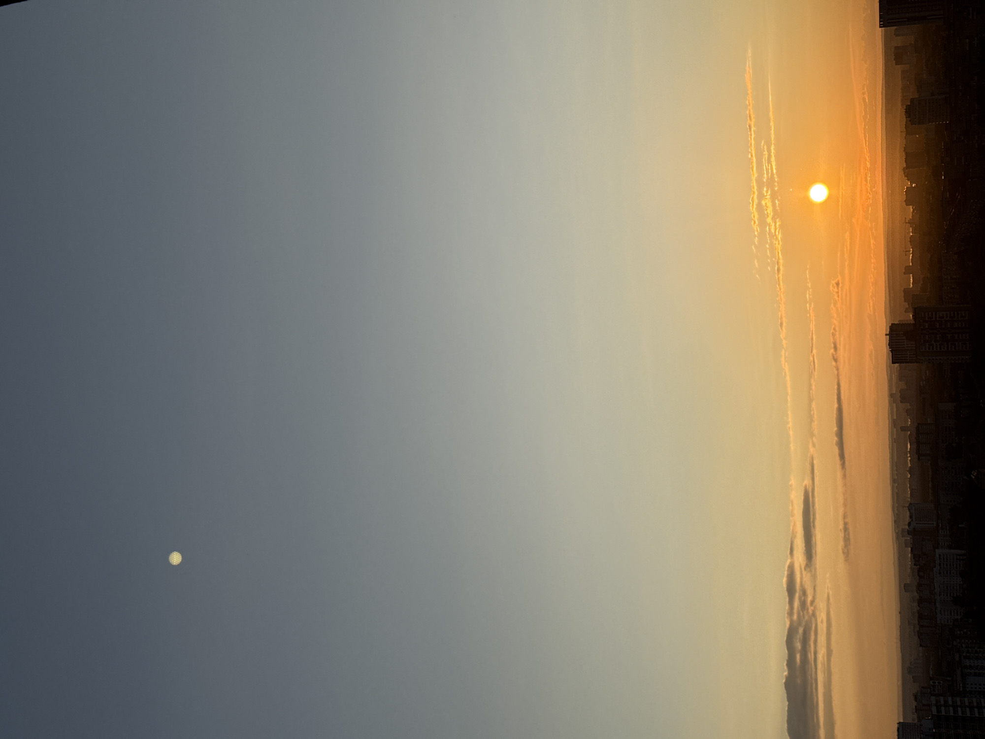 The image, best viewed rotated 90 degrees counter-clockwise, depicts a wide panoramic view of the sky over a distant city horizon, likely during sunrise or sunset. The sky presents a striking gradient, transitioning from a deep blue-grey on the left to vibrant orange and yellow hues on the right, illuminated by a bright sun.
On the right side of the frame, the sun appears as a brilliant, glowing orange orb, casting its warm light across the atmosphere. Several elongated clouds, tinged with orange and yellow, stretch across the sky near the sun, catching its rays. Along the horizon line beneath the sun, the faint silhouettes of distant city buildings and structures are visible, appearing somewhat hazy due to atmospheric conditions or distance.
In contrast, on the far left of the image, set against the darker blue-grey sky, a distinct bright, circular object is present. Upon closer inspection, this object has a noticeable patterned or grid-like surface, suggesting it may be a highly reflective artificial light source or a unique atmospheric phenomenon rather than the natural moon.
The scene captures a moment of transition, highlighting the vastness of the sky and the atmospheric effects of the sun's low position, overlooking an urban expanse in Perm, Russia. No discernible text is visible in the image.