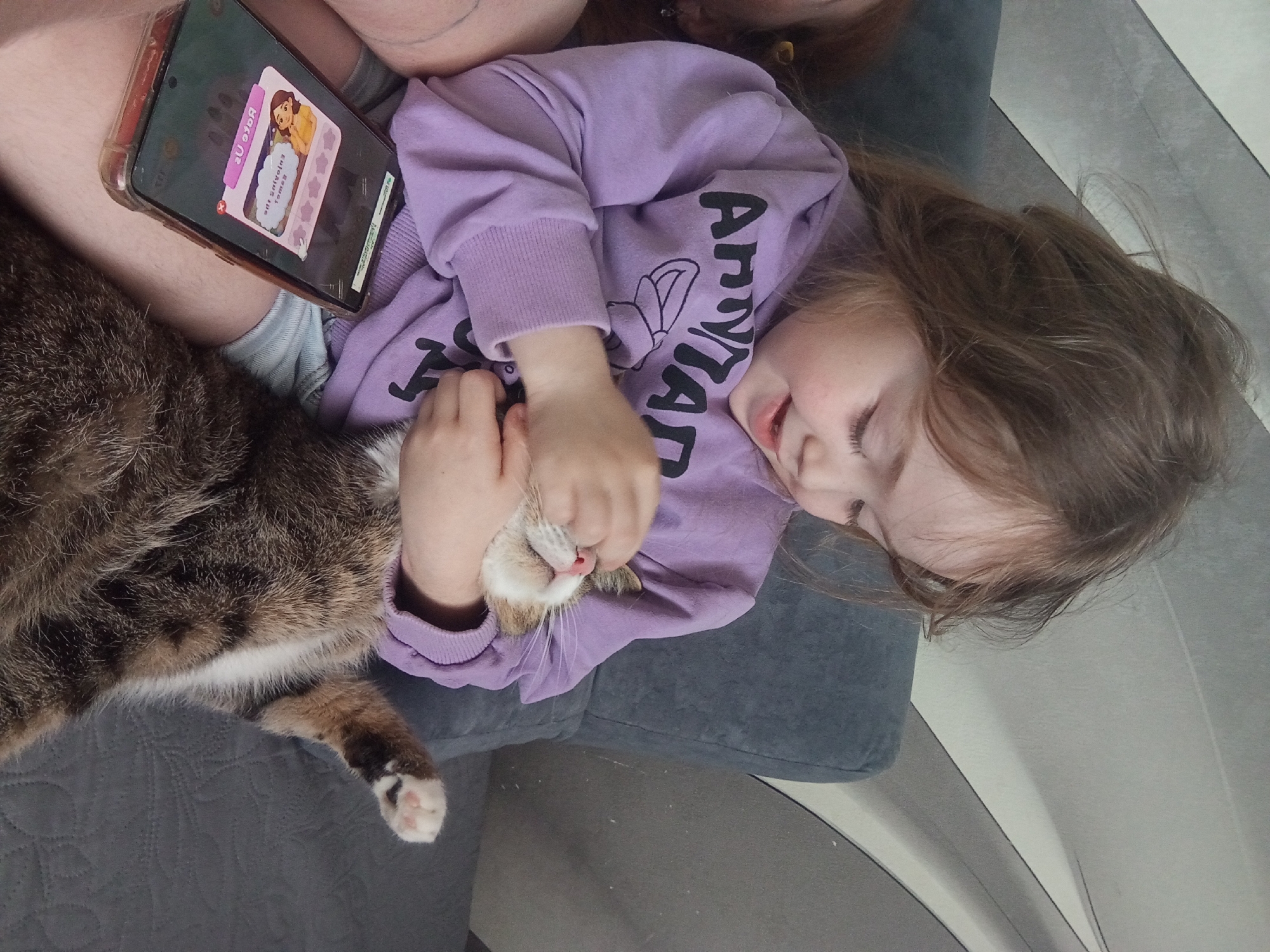 A young girl and a tabby cat are captured in a relaxed, intimate moment indoors. The girl, with light brown hair, is lying on her back, wearing a purple long-sleeved top with "APPYLAND" printed in black on the front. Her eyes are mostly closed or cast downwards, and a soft smile plays on her lips, indicating contentment. Her hands are gently cradling the face of the cat, which is also lying on its back, its eyes closed and a small pink nose visible, suggesting it's enjoying the interaction.
The cat is a domestic tabby, with a mix of brown, black, and white fur, and a white chest and paws, one of which shows delicate pink toe beans. They are resting together on a grey, textured upholstered surface, likely a couch or bed.
Above the girl's legs and beside her, a smartphone in a clear case is visible, resting on what appears to be an adult's thigh. The phone screen is active, displaying a colorful game or application. Text on the screen includes "Вверх" (Up) at the top, and below an animated character, some upside-down Russian text that appears to be "игрушечную коробку для хранения" (toy storage box). The scene conveys a peaceful and affectionate interaction between the child and her pet, possibly during a quiet moment at home in Yurgamysh, Russia.