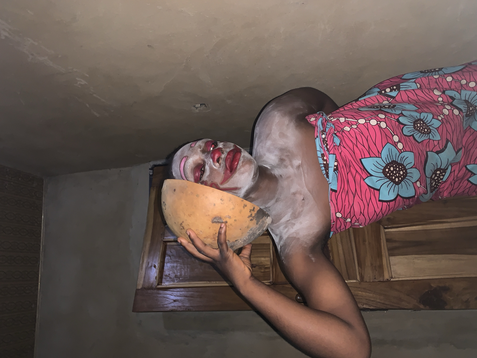 In the city of Agban, Nigeria, a person with white face paint and red clown makeup is posing. The makeup includes red lipstick and rosy cheeks. The individual is wearing a pink and blue floral wrap skirt. They are holding a hollowed-out gourd to the left side of their face. The background consists of a wooden door and a textured wall, suggesting an indoor setting.
