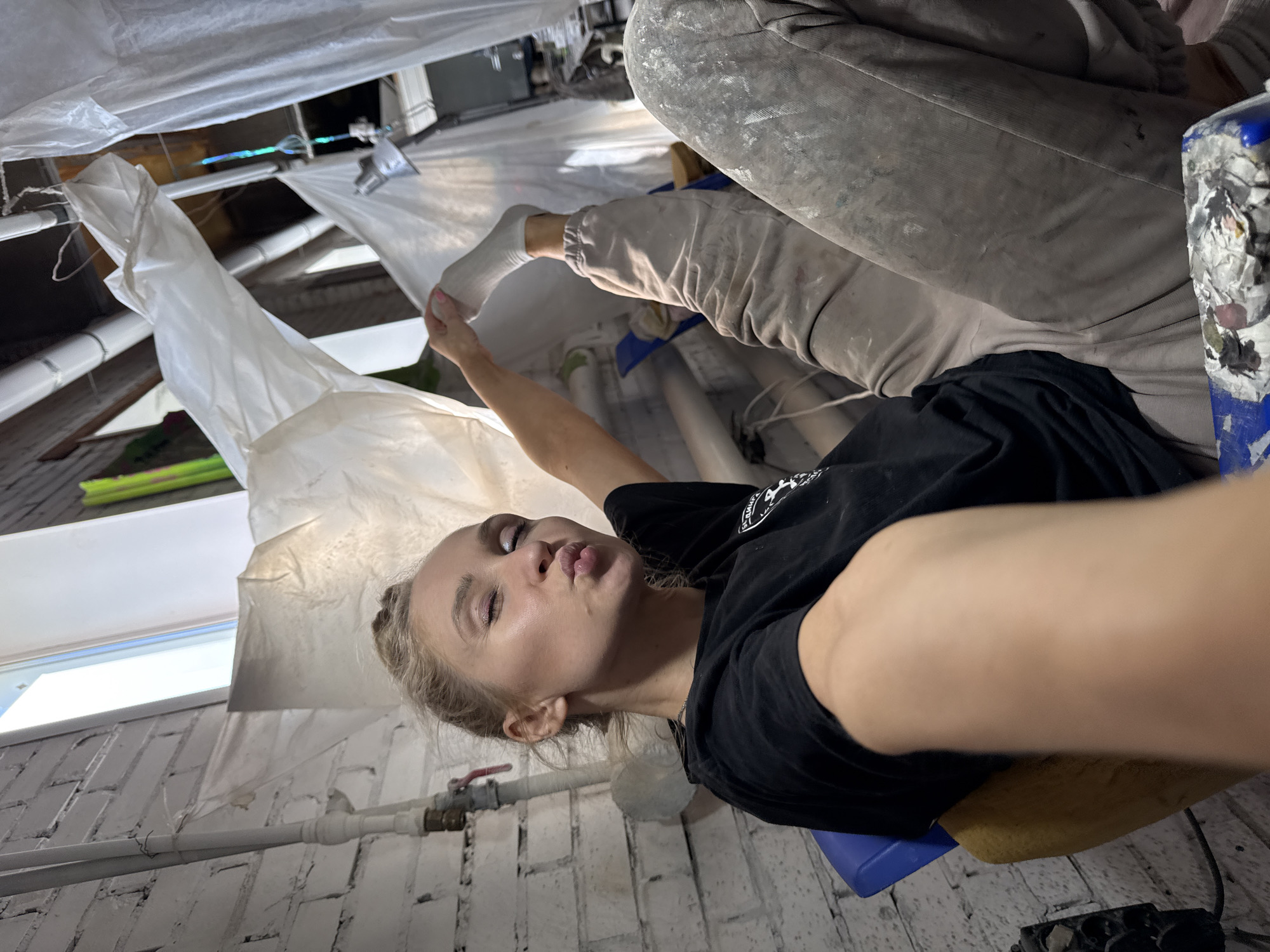A blonde woman is in the foreground, wearing a black t-shirt and looking towards the camera with pursed lips. Her left arm is outstretched as if taking a selfie. She is sitting on a blue surface, likely a chair.
The background shows a room covered with plastic sheeting. Visible are walls painted white with brick-like patterns and pipes running along them. A pair of legs, clad in paint-splattered grey pants and a white sock, are in the upper right quadrant, partially obscured by the woman's arm and the plastic sheeting. The room appears to be under renovation.
The city of Chaikovsky, Russia is not evident in the image.