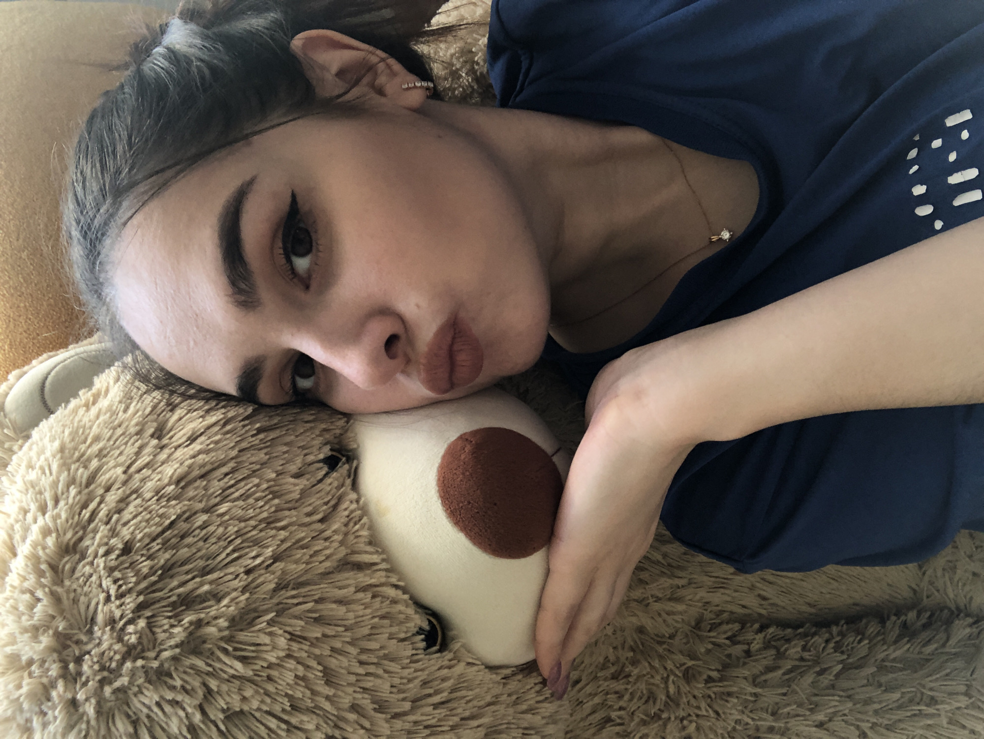 A young woman is lying down with her head resting on a tan-colored teddy bear. She has dark hair pulled back and is wearing a dark blue shirt and a necklace with a diamond pendant. She has a light complexion with dark eyebrows and eye makeup. Her left arm is extended, her hand placed on the teddy bear's face. The background and surrounding surfaces are textured and appear to be plush or furry. The location is in the city of Noviy, Russia.
