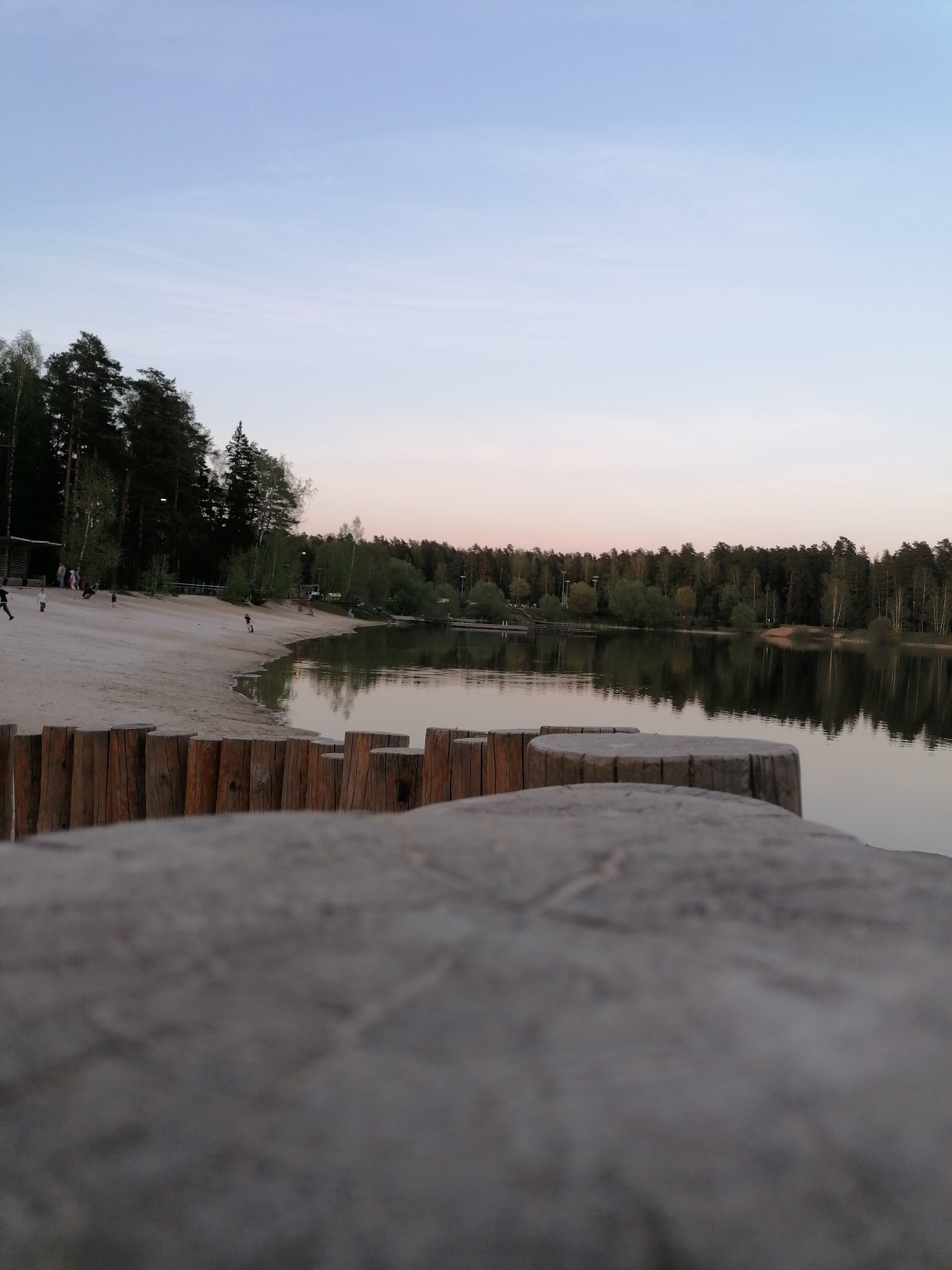 The image captures a lakeside scene at dusk. A sandy beach is visible, dotted with sparse vegetation and trees lining the shore. Several individuals are present on the beach, though their activities are unclear. A wooden barrier constructed from upright logs separates the foreground from the water's edge. The lake's surface is calm, reflecting the surrounding trees and sky. The sky transitions from light blue to a faint pinkish hue, indicating the approach of night. The scene appears to be in a park or recreational area in the city of Unknown, Unknown.
