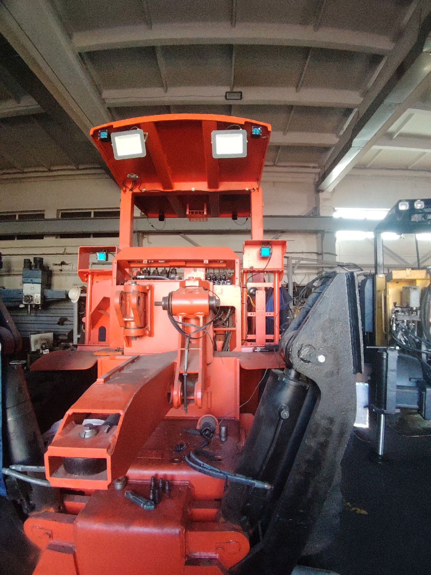 The image depicts a large, orange industrial machine, possibly a forestry or construction vehicle, situated indoors within a workshop or factory setting. The machine features prominent hydraulic components, a heavy-duty articulated arm, and multiple lights mounted on an overhead canopy. The environment shows industrial shelving, exposed ceiling beams and ductwork, and other machinery in the background, suggesting an active manufacturing or repair facility. The overall impression is one of heavy machinery in a utilitarian industrial space. There are no people visible in the image. The location is identified as Oskemen, Kazakhstan.
