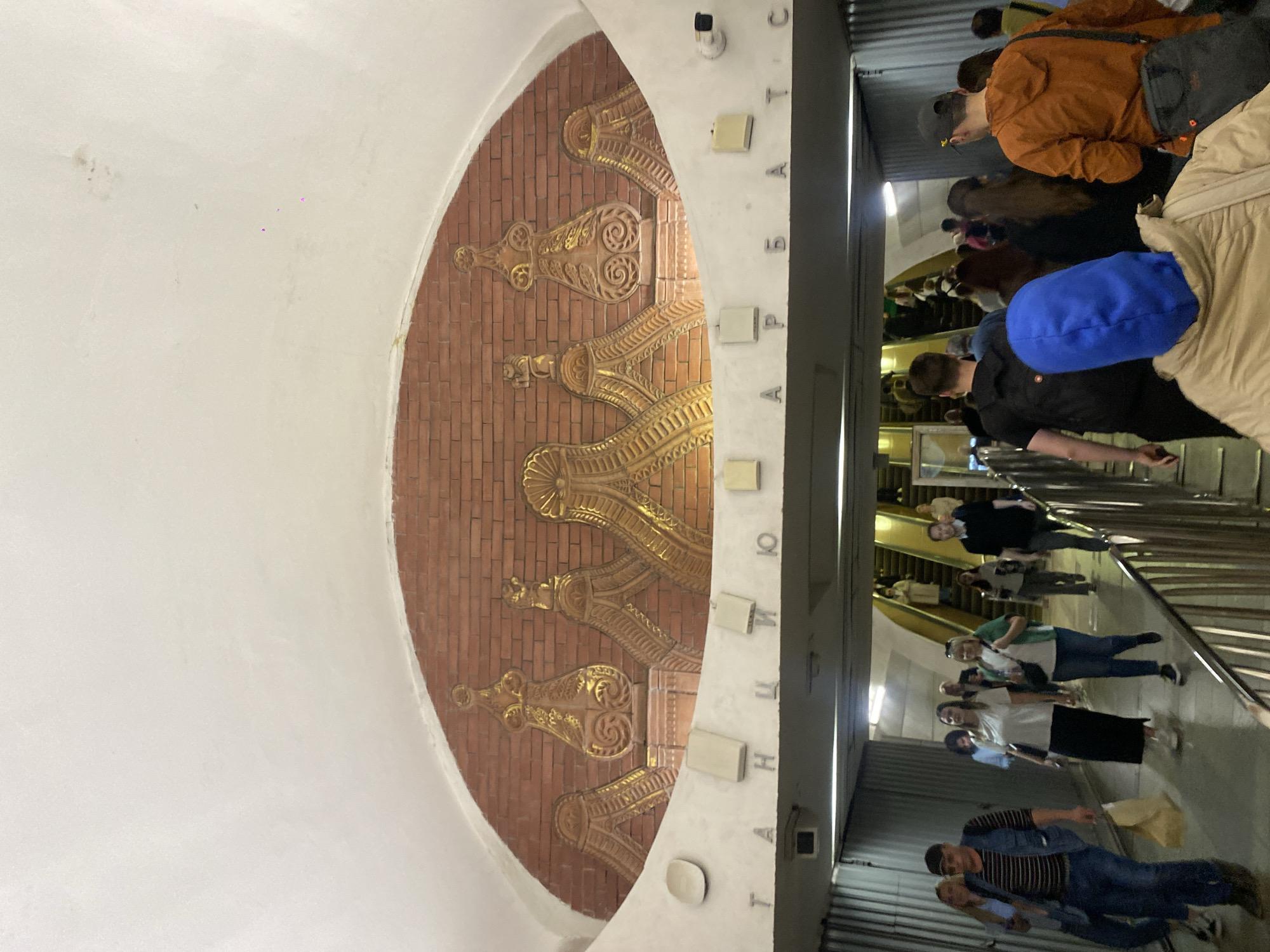 The image shows a wall decoration in the Arbatskaya metro station in Moscow, Russia, featuring a brick background with gold-colored decorative elements. The wall is arched and flanked by a white wall. In the background, escalators transport people. A diverse group of people is visible.