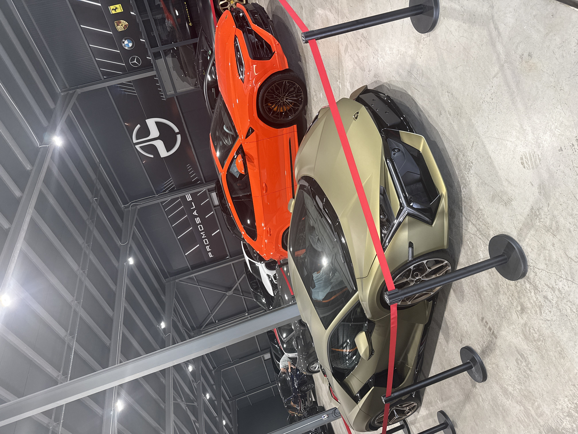 The image depicts the interior of a high-end car showroom, likely in Plovdiv, Bulgaria, as suggested by the mention of "PROMOSALE" which is a business located there. The scene is dominated by several luxury vehicles. In the foreground, a matte gold Lamborghini Aventador is prominently displayed, with its sharp lines and aggressive styling visible. A red velvet rope barrier with black stanchions delineates the space around the car. Behind the Aventador, an orange Lamborghini Urus SUV is parked. Further in the background, other dark-colored luxury cars, including what appears to be a dark blue Audi and a black Porsche, are visible. The showroom features a modern industrial aesthetic with a high, exposed ceiling with metal beams and track lighting. A large black wall in the background is adorned with logos of luxury car brands such as Mercedes-Benz, BMW, and Porsche, as well as a stylized "PS" logo and the text "PROMOSALE". The overall impression is one of wealth and exclusivity.