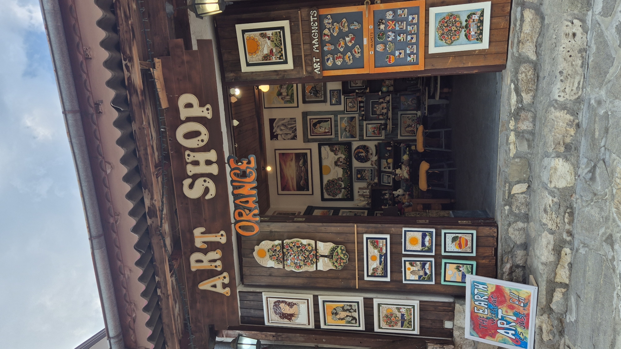 The image shows the exterior of an art shop named "ORANGE ART SHOP" located in the city of Nesebar, Bulgaria. The shop has a wooden facade with a traditional tiled roof. Various artworks, including paintings, mosaics, and magnets, are displayed on the exterior walls and inside the open doorway. A sign at the entrance reads "ART SHOP ORANGE". A white sign on the steps in front of the shop displays the text "THE EARTH without ART is just EH!". The scene appears to be an outdoor market or street with a stone-paved walkway and steps. No people are visible in the image.