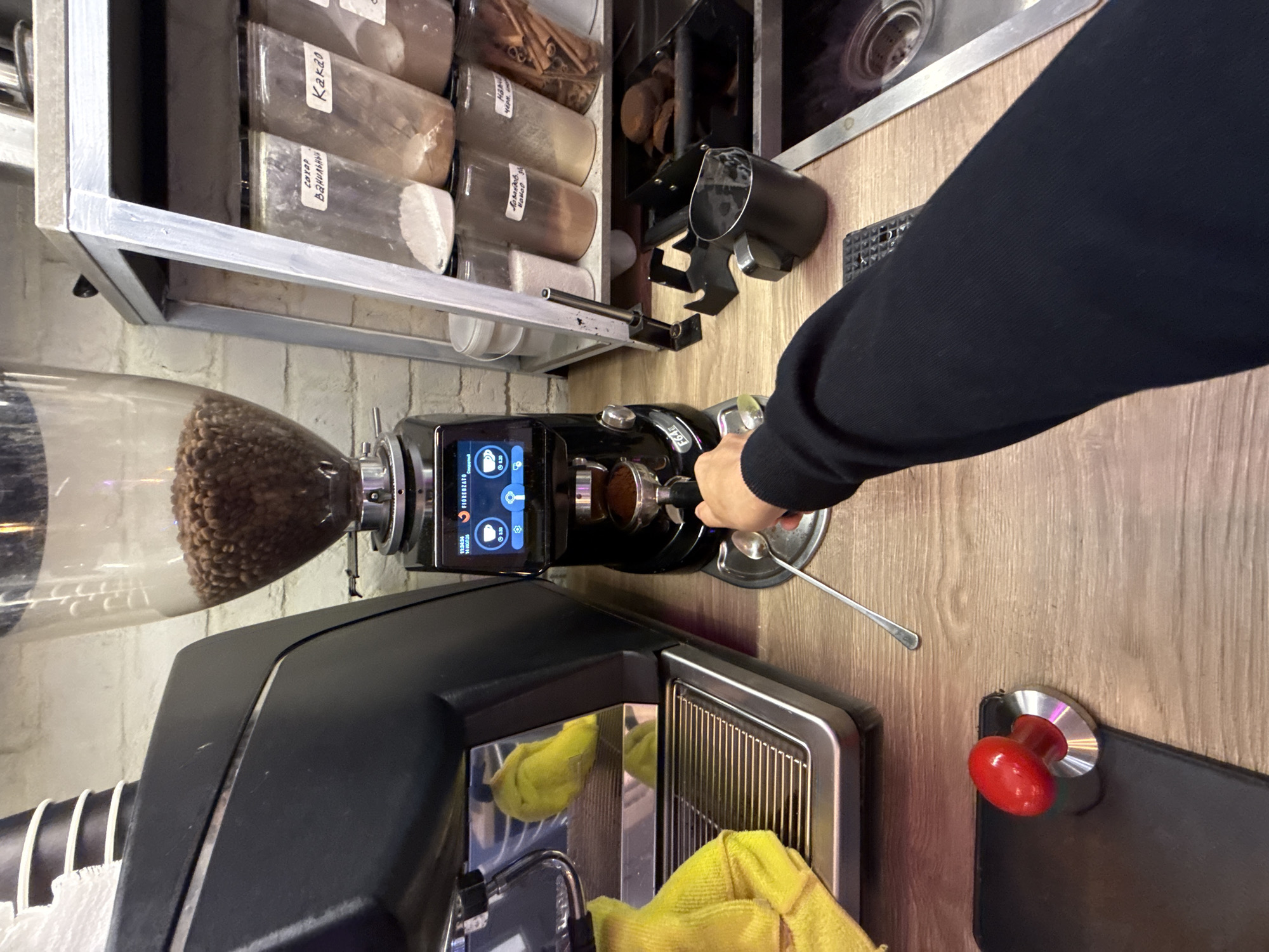 This image depicts a close-up, point-of-view shot of a person's arm and hand operating a coffee grinder and espresso machine in what appears to be a commercial setting. The scene is set on a wooden countertop. In the foreground, a red-handled tamper rests on a black surface. The main subject is a black coffee grinder with a clear hopper filled with coffee beans, a digital display showing "FIORENZATO," and a portafilter containing ground coffee being held by the person's hand. A silver spoon lies on the counter near the portafilter. Behind the grinder, an espresso machine with a yellow cloth draped over its side is visible. To the right, a metal shelf holds several clear plastic containers with various powdery ingredients labeled in Cyrillic script, suggesting a location where these ingredients are stored and used, possibly for making coffee drinks. One container appears to hold cinnamon sticks. A black pitcher and a metal tray with what look like cookies or coffee pucks are also present. The background shows a white brick wall. There is no visible indication of the specific location within Moscow, Russia.