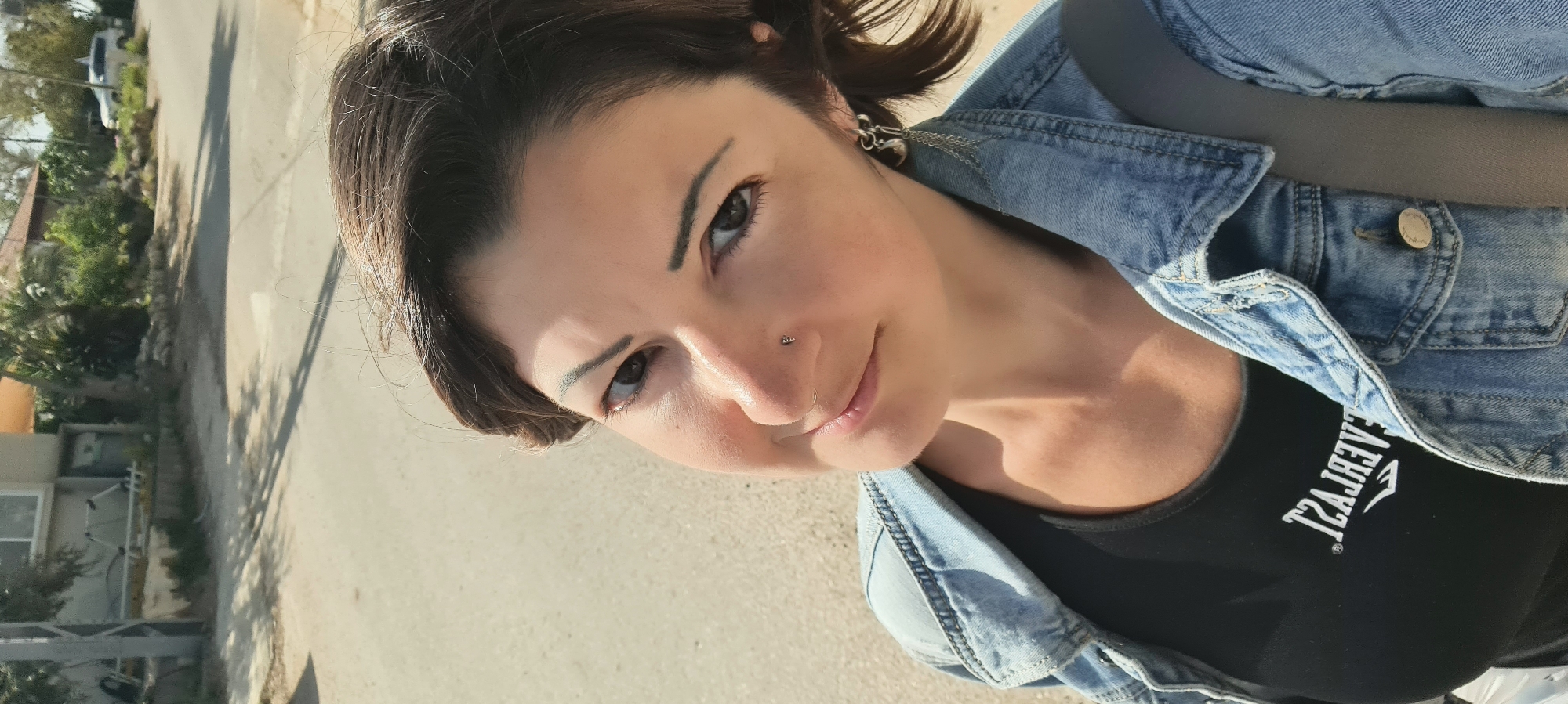 A woman is featured prominently in an outdoor portrait, looking directly at the camera with a gentle smile. She has short, dark hair, dark eyes, and a fair complexion. Notable facial features include defined eyebrows, a small stud piercing on the side of her nose, and a nose ring in her nostril. Her visible ear has an earring featuring a chain and a heart-shaped pendant.
She is wearing a light blue denim jacket over a black top. The black top displays white text, which, due to the image's orientation or a selfie camera flip, appears reversed but is clearly identifiable as "EVERLAST."
The background shows an outdoor setting with strong sunlight casting shadows on a paved surface, likely a road or pathway. In the mid-ground and distance, there are residential buildings with light-colored walls and tiled roofs, partially obscured by green foliage, including trees and bushes. The bright lighting suggests it is daytime with clear weather. The image perspective, looking slightly upwards at the woman, suggests it might be a selfie taken outdoors in the Lachish Regional Council, Israel.