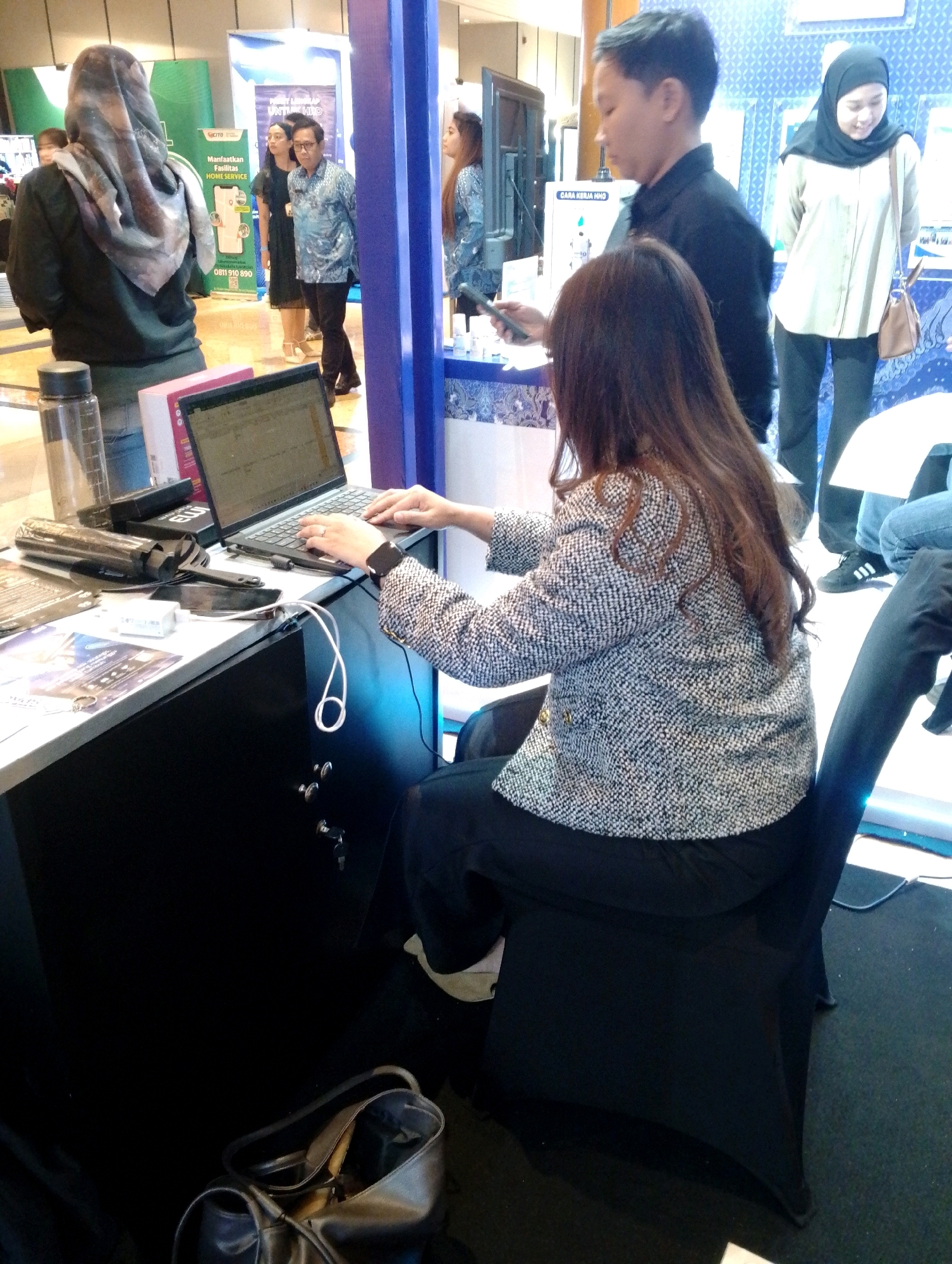 The image captures a busy indoor scene, possibly at an exhibition or event, in an unknown city. In the foreground, a woman with long brown hair is seated at a desk, engrossed in typing on a laptop. She is wearing a patterned jacket and dark pants, and a smartwatch is visible on her left wrist. The desk is cluttered with various items, including a water bottle, a charging cable, a smartphone, and some papers. To the left of the desk, a dark handbag rests on the floor.
Behind the woman at the desk, a man in a black shirt is standing, looking towards the right side of the frame. He is holding a smartphone in his hand. To the right of him, a woman wearing a black hijab and a light-colored shirt is standing and looking downwards, with a brown handbag slung over her shoulder.
Further in the background, other individuals are visible, including a woman in a headscarf to the far left, and several other people standing and walking in the vicinity. The environment appears to be an indoor exhibition hall with booths and displays set up. Blue and white backdrops are visible, suggesting a professional or commercial setting. The lighting is a mix of overhead illumination and possibly some spotlighting.
