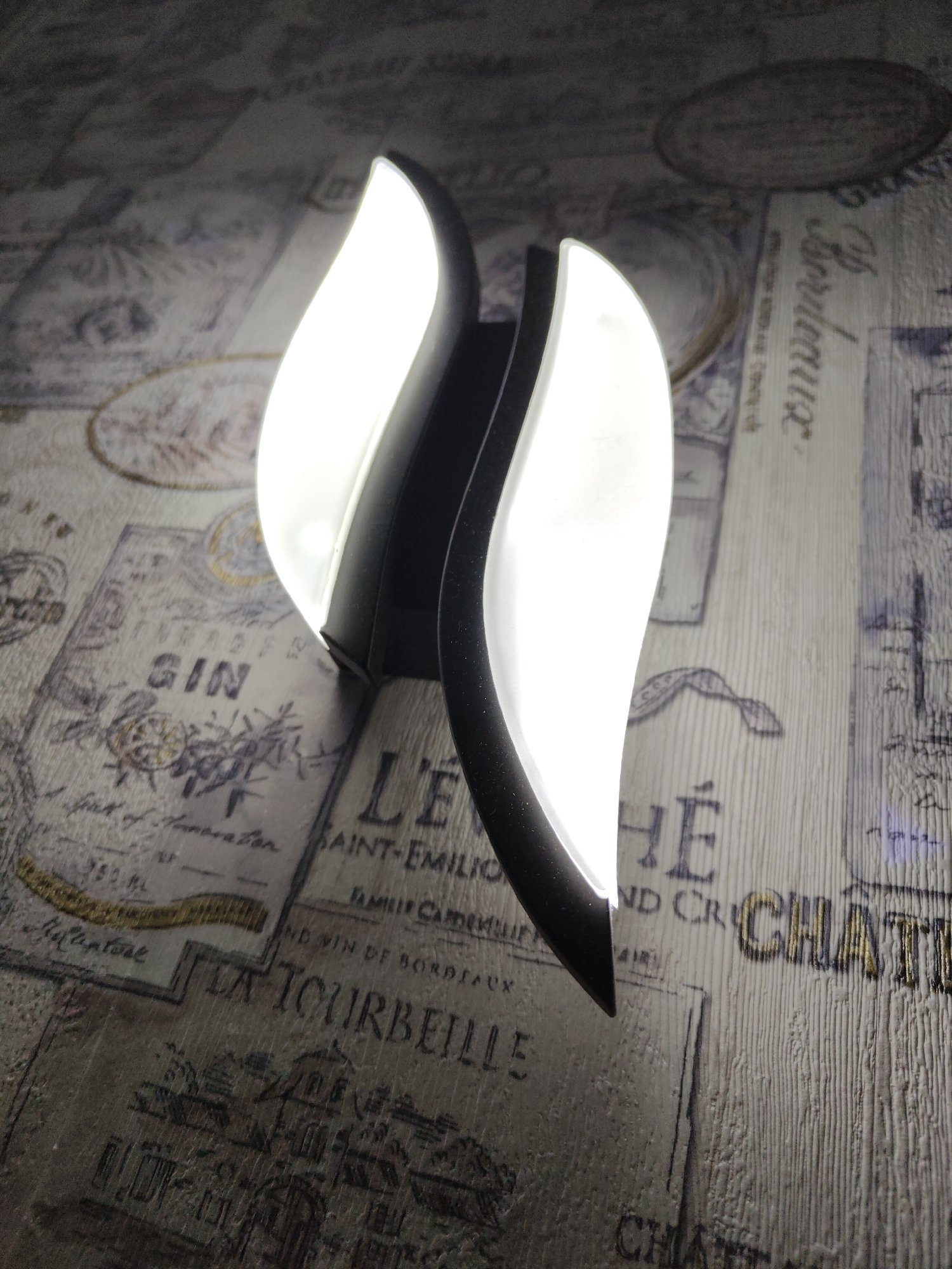 Here is a description of the image.
The image shows a modern wall sconce with two curved, illuminated white panels encased in a matte black frame. The sconce is mounted on a wall covered in patterned wallpaper that features vintage-style designs with what appears to be writing in French. The wallpaper's color palette includes shades of gray, cream, and touches of gold, giving it an aged, yet elegant, look. Inscriptions on the wallpaper include the words "GIN," "SAINT-EMILIO," "LA TOURBEILLE," "Bordeaux," and "CHATI" suggesting a theme related to wine or alcoholic beverages. There is no information in the image to place it in the city of Ufa, Russia.