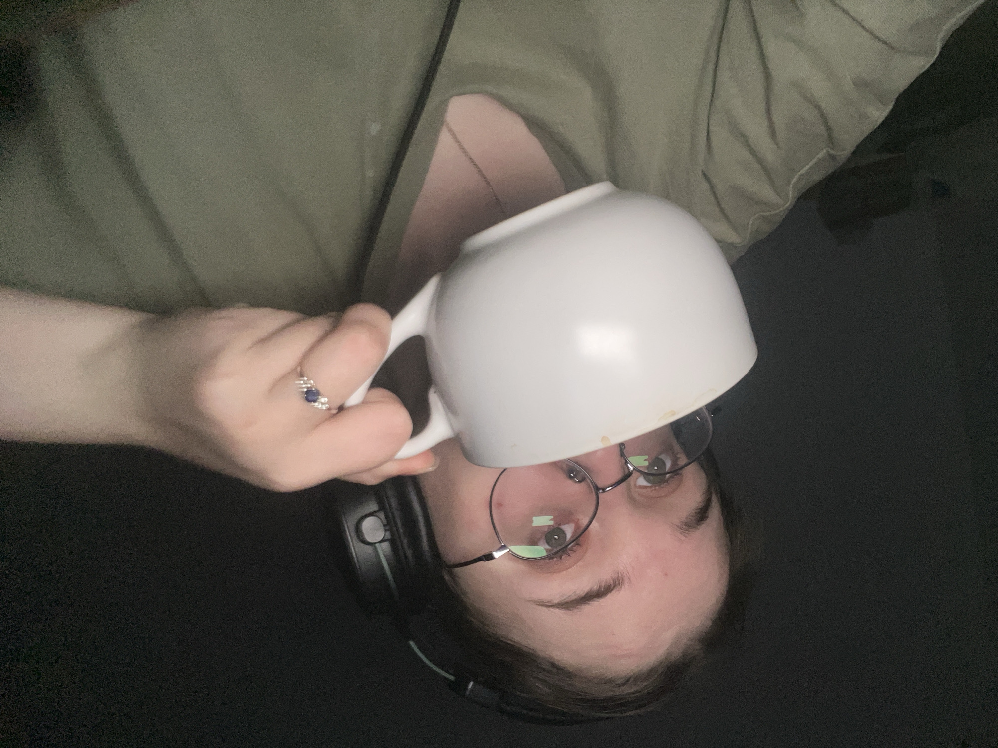 A person with fair skin is holding a white mug, seemingly drinking from it. They are wearing round eyeglasses with a thin metal frame and headphones. A ring with a dark blue stone is on their finger. The person is wearing a light green shirt with a chain necklace visible at the neckline. The background is dark and out of focus. There is no indication of the location being Pushchino, Russia.