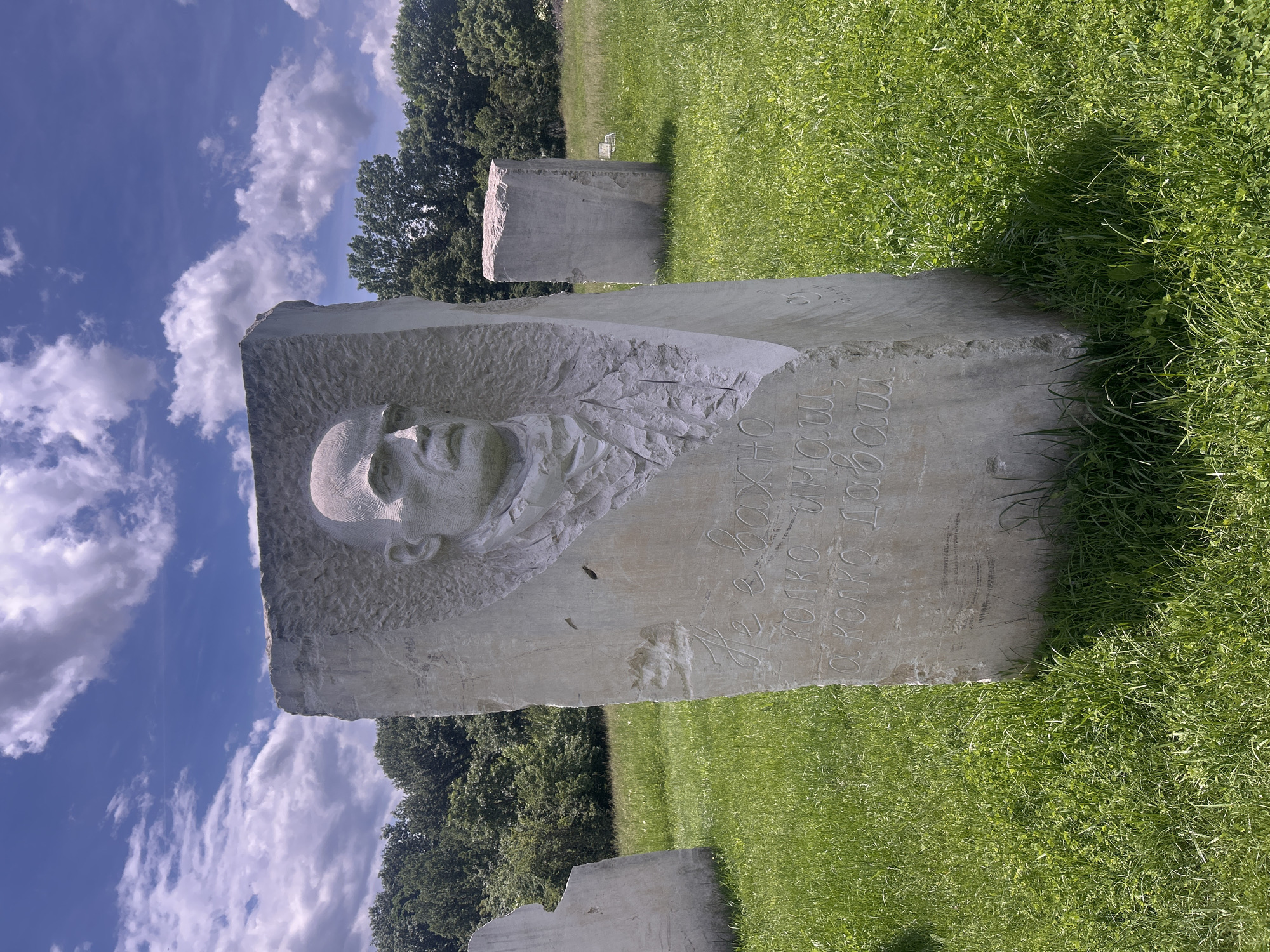 The image displays an outdoor scene featuring a large, upright grey stone sculpture situated in a vibrant green grassy field under a bright blue sky. The most prominent subject is the front face of this main sculpture, which features a detailed relief carving of a bald man's head and upper chest. The man has a solemn or determined expression.
Adjacent to the carved bust, Cyrillic text is etched into the stone. The text reads: "Не важно сколько им а сколько даш". This translates to "It's not important how much they have, but how much you give."
In the background, several other similar, roughly shaped or less detailed stone blocks are scattered across the expansive green lawn, suggesting an outdoor sculpture park or a memorial site. A dense line of green trees forms the horizon beyond the field. The sky is a clear, brilliant blue, adorned with scattered fluffy white cumulus clouds. The bright illumination and visible shadows indicate a sunny day, likely midday or early afternoon.
The scene, located in Gardevtsi, Bulgaria, captures a serene yet monumental setting, combining artistic expression with a philosophical message inscribed on the stone.