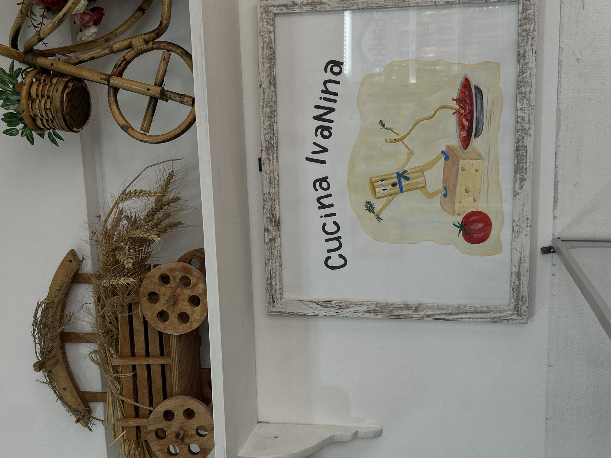 Image features a close-up of a wall decorated with various objects. On the left, there is a decorative wooden bicycle and wagon, adorned with artificial flowers, greenery, and dried wheat. To the right, a framed picture hangs on the wall. The picture contains the words "Cucina Ivanina" in Italian and depicts a cartoon image related to Italian food, with spaghetti, cheese, and a tomato. The wall itself appears to be white. The location is in the city of Sofia, Bulgaria.
