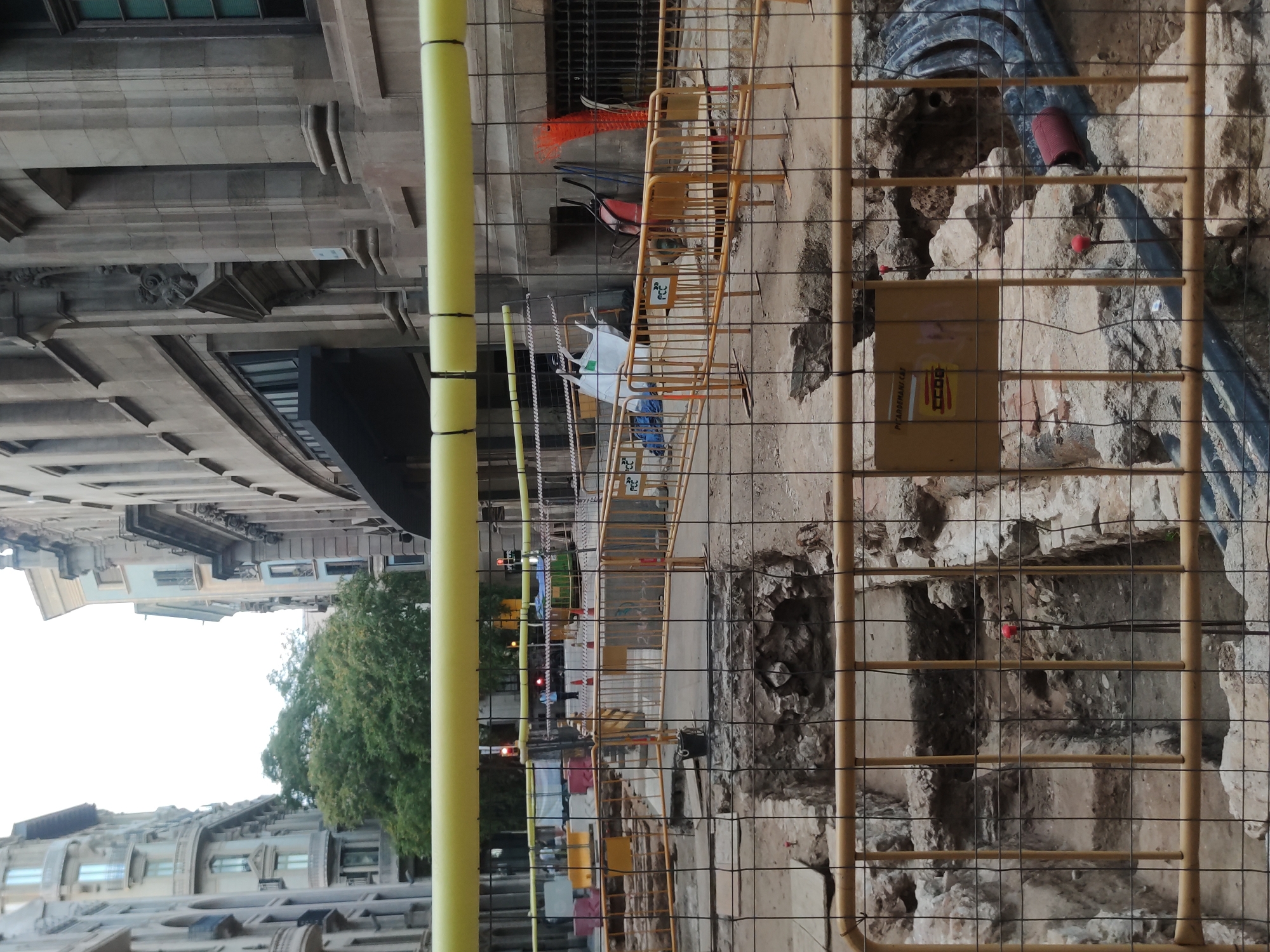 This is a photograph of an archaeological excavation site in Barcelona, Spain. The site is located between tall, old buildings, suggesting an urban environment. The excavation itself reveals stone structures and earth, with some debris and construction materials visible. The area is cordoned off with yellow metal barriers and black wire fencing, indicating ongoing work and restricted access.
In the background, a yellow tubular structure spans horizontally across the image, possibly a part of temporary scaffolding or a utility line. Beyond the barriers, there are signs of a street with traffic lights and vehicles, as well as some people in the distance. Building facades with architectural details are visible on either side of the street, typical of Barcelona's cityscape. The overall impression is one of urban development or preservation work happening in a historical part of the city.