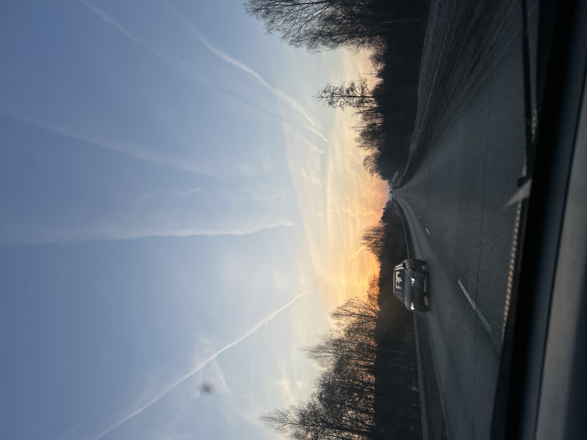 The image shows a road near Garevaya, Russia, viewed from inside a vehicle. A car is on the road in the opposite lane. The sky shows contrails and an orange glow near the horizon, likely indicating sunrise or sunset. Trees line both sides of the road. The road surface is dark and appears to be asphalt.