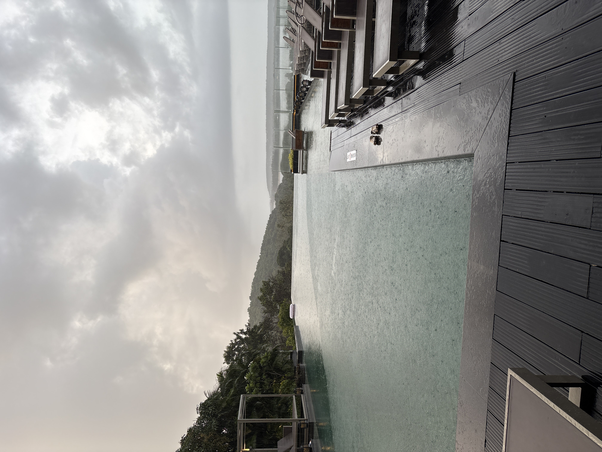 The image depicts an outdoor swimming pool area in Vagator, India, during a rain shower. The infinity pool dominates the foreground, with visible raindrops creating ripples on its surface. A pink inflatable ring floats near the edge of the pool. To the left, lounge chairs are visible under a covered structure. The surrounding area features lush green trees and hills in the background, with a body of water visible in the distance. The sky is overcast with heavy clouds, suggesting a gloomy or stormy weather condition. A wooden deck with multiple lounge chairs is situated beside the pool, with some chairs appearing wet from the rain. A pair of sandals are placed on the deck near the edge of the pool. The overall atmosphere is serene yet melancholic due to the weather.