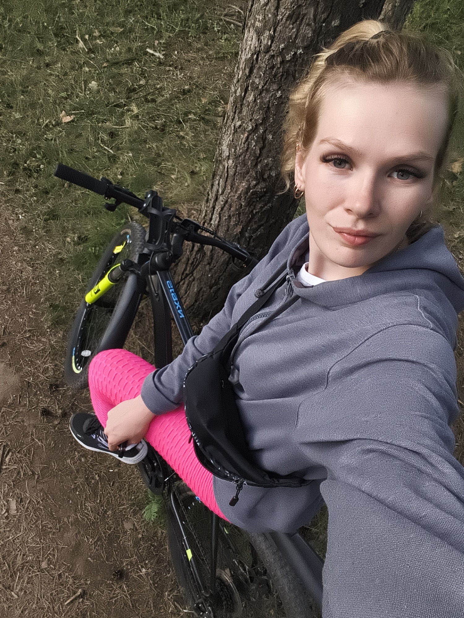 An overhead, eye-level, medium shot captures a woman resting with her bicycle against a tree in a grassy area. The woman, with blonde hair pulled back in a ponytail, is wearing a gray hooded sweatshirt over a white t-shirt, bright pink textured leggings, and black sneakers. She has a black fanny pack slung across her chest. Her right leg is bent at the knee, with her foot resting on the bicycle's frame. The bicycle, dark-colored with yellow accents on the front fork, is positioned vertically next to the tree trunk. The ground is covered with dry grass, twigs, and pine cones, suggesting an outdoor, possibly wooded, location. The lighting is natural, creating soft shadows. The image was taken in Русский Вожой, Russia.