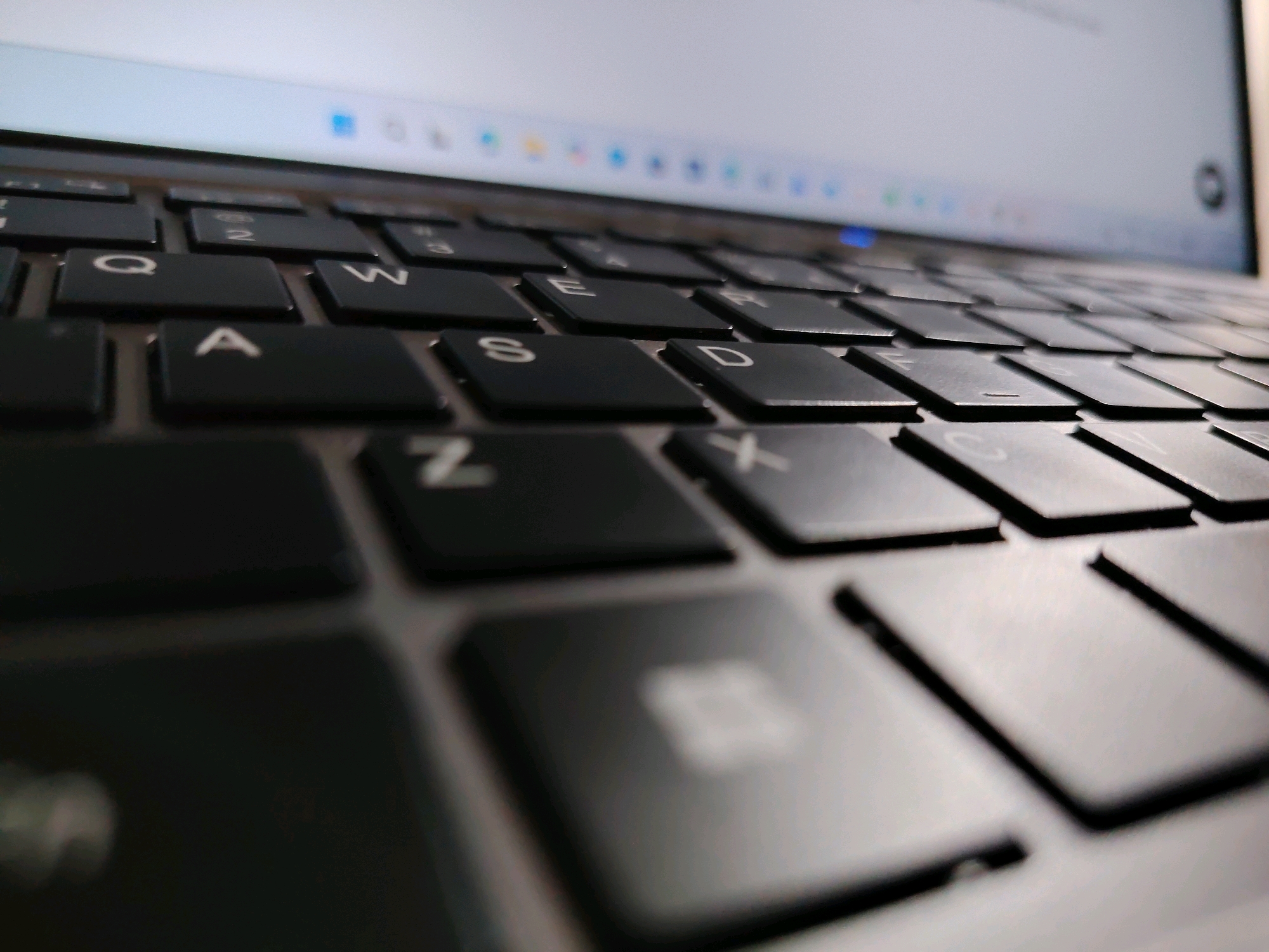 The image is a close-up, angled shot of a laptop keyboard. The keys are black with white lettering. The focus is on the keys in the middle and right side of the keyboard, showing letters like 'Q', 'W', 'E', 'A', 'S', 'D', 'Z', 'X', and other partial characters. The laptop screen is visible in the background, out of focus, showing a web browser interface with icons and a blue address bar. The lighting suggests an indoor environment, likely in the daytime, with a soft glow reflecting on the keyboard. There are no people or specific locations identifiable within the image, and therefore no connection to Davao City, Philippines can be made.
