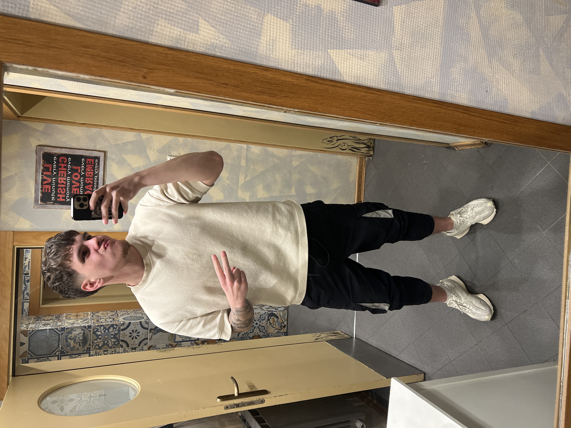 This image depicts a young male taking a mirror selfie in an interior space in Sofia, Bulgaria, likely a hallway or a private residence.
The subject is a young male with short, textured brown hair, a faded haircut on the sides, and light facial stubble on his chin. He wears a cream-colored short-sleeved crew-neck t-shirt, black cargo pants with light grey accents on the side pockets, and cream-colored athletic shoes. His left forearm displays visible tattoos. He is holding a black smartphone with his right hand to take the photo, while his left hand is raised, forming a "peace" sign (V-sign). His lips are pursed, and his gaze is directed towards the phone/mirror.
The environment features a dark grey tiled floor. To the subject's left, a light-colored wooden door with a round, frosted glass panel (patterned with flowers) and a gold-colored handle is partially open. A grey radiator is visible below the door handle. The wall behind the door features an arrangement of decorative ceramic tiles in blue, white, and dark patterns, some depicting figures. To the subject's right, a full-length mirror with a prominent light brown wooden frame reflects the scene. The wall directly adjacent to and reflected in the mirror has a light-colored, textured wallpaper with a subtle geometric pattern. A rectangular sign with reversed white text on a red background is visible above the subject's right shoulder in the reflection; it reads "LIVE every minute, CHERISH every memory, LOVE beyond words, EMBRACE every possibility." A white, flat-topped counter or cabinet is visible in the bottom left foreground.