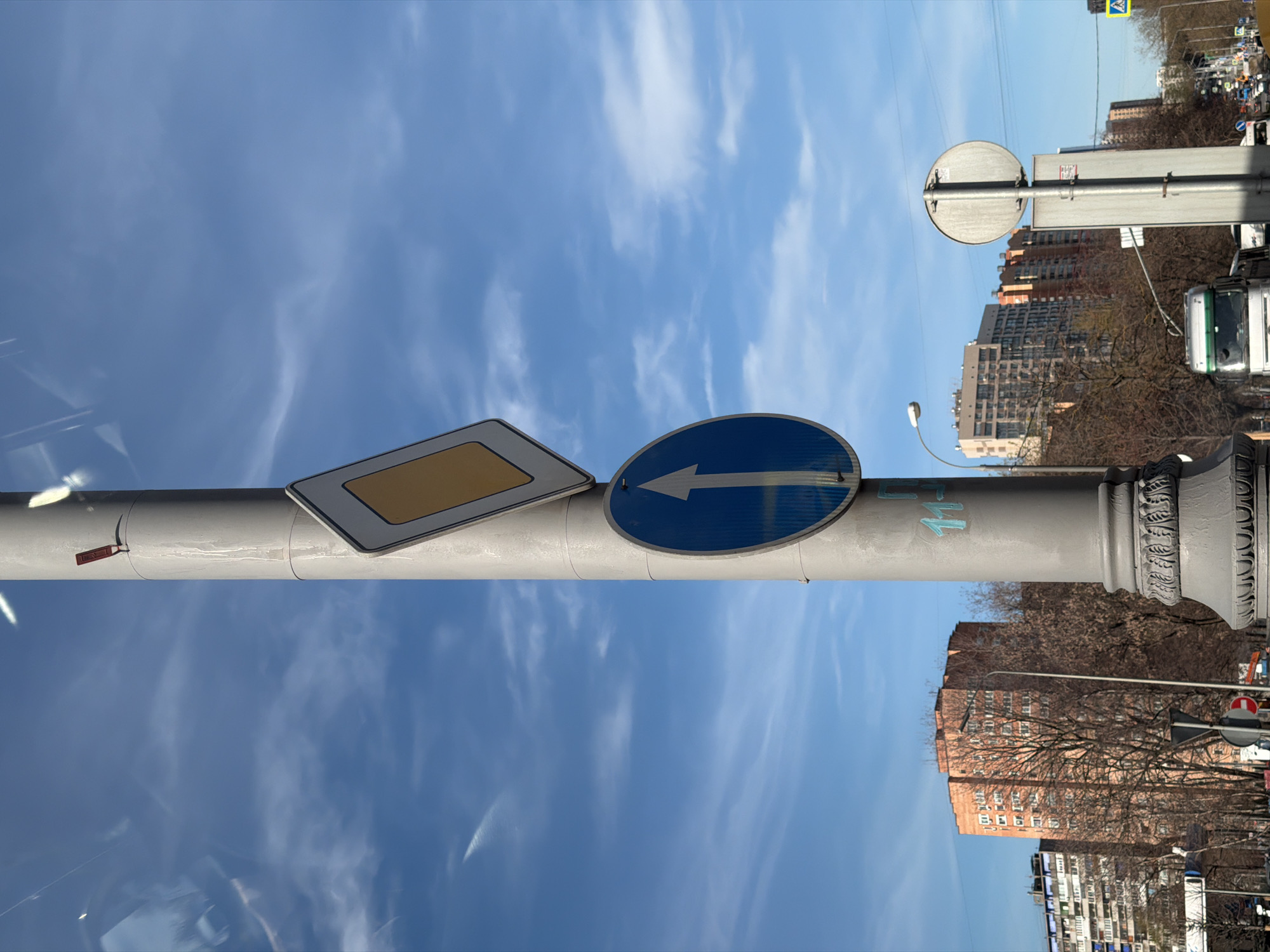 A street scene in Moscow, Russia, captured during the daytime under a bright blue sky with scattered wispy clouds. The central focus is a tall, grey pole positioned vertically in the frame (if rotated correctly). Attached to this pole are two prominent road signs. On the left, there is a rectangular yellow sign with a thick white border, characteristic of a "priority road" sign (Главная дорога). To its right, a circular blue sign displays a white arrow pointing left, indicating a mandatory direction. Faint blue graffiti or writing, possibly Cyrillic characters like 'И' and 'Ч', can be seen on the pole below the signs.
In the background, multi-story residential buildings with varying shades of brown and beige facades are visible. Bare-branched trees, suggesting late winter or early spring, stand between the buildings and the pole. Street furniture, including another sign pole with a circular sign (possibly a "no entry" or "stop" sign) and a lamppost, can be discerned in the middle ground. Further in the distance, above the buildings, a blue square sign with a white triangle depicting a pedestrian, indicating a pedestrian crossing, is partially visible. A white and green truck or bus is partly seen on the far right side of the image. Reflections on a surface, likely a vehicle window from which the photograph was taken, are present in various parts of the image, particularly in the lower left.