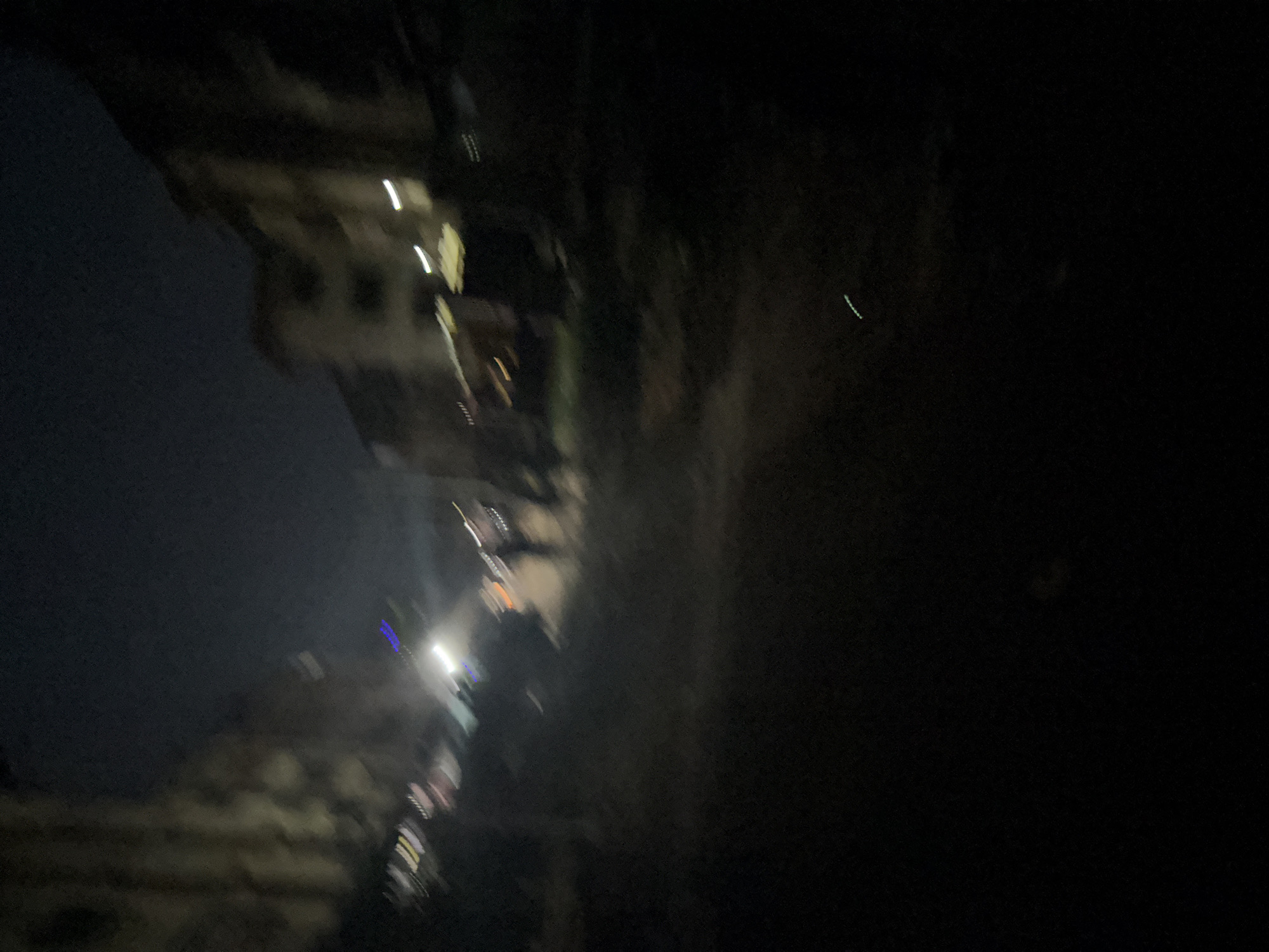 The image depicts a blurry nighttime scene of a street in Onitsha, Nigeria. Buildings line the sides of the street, with some windows illuminated by artificial light. Several figures are visible, appearing as indistinct silhouettes moving along the street. Streaks of light suggest movement, possibly from vehicles or streetlights. The overall atmosphere is dark and somewhat disorienting due to the motion blur, characteristic of a low-light environment where the camera was likely in motion.