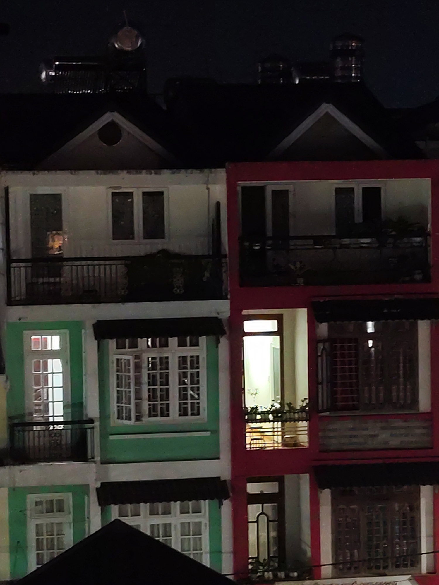 At night, the image shows two buildings of differing colors side-by-side in what is believed to be Da Lat, Vietnam, evidenced by the architectural style. The building on the left is painted white and green, while the building on the right is painted red. Both buildings appear to be multi-story with multiple windows, some with open or closed shutters, and balconies with railings. There are lights visible inside some of the rooms. At the top of the image, the dark roofs of more buildings are visible, with some cylindrical structures that appear to be water heaters. The sky is dark.