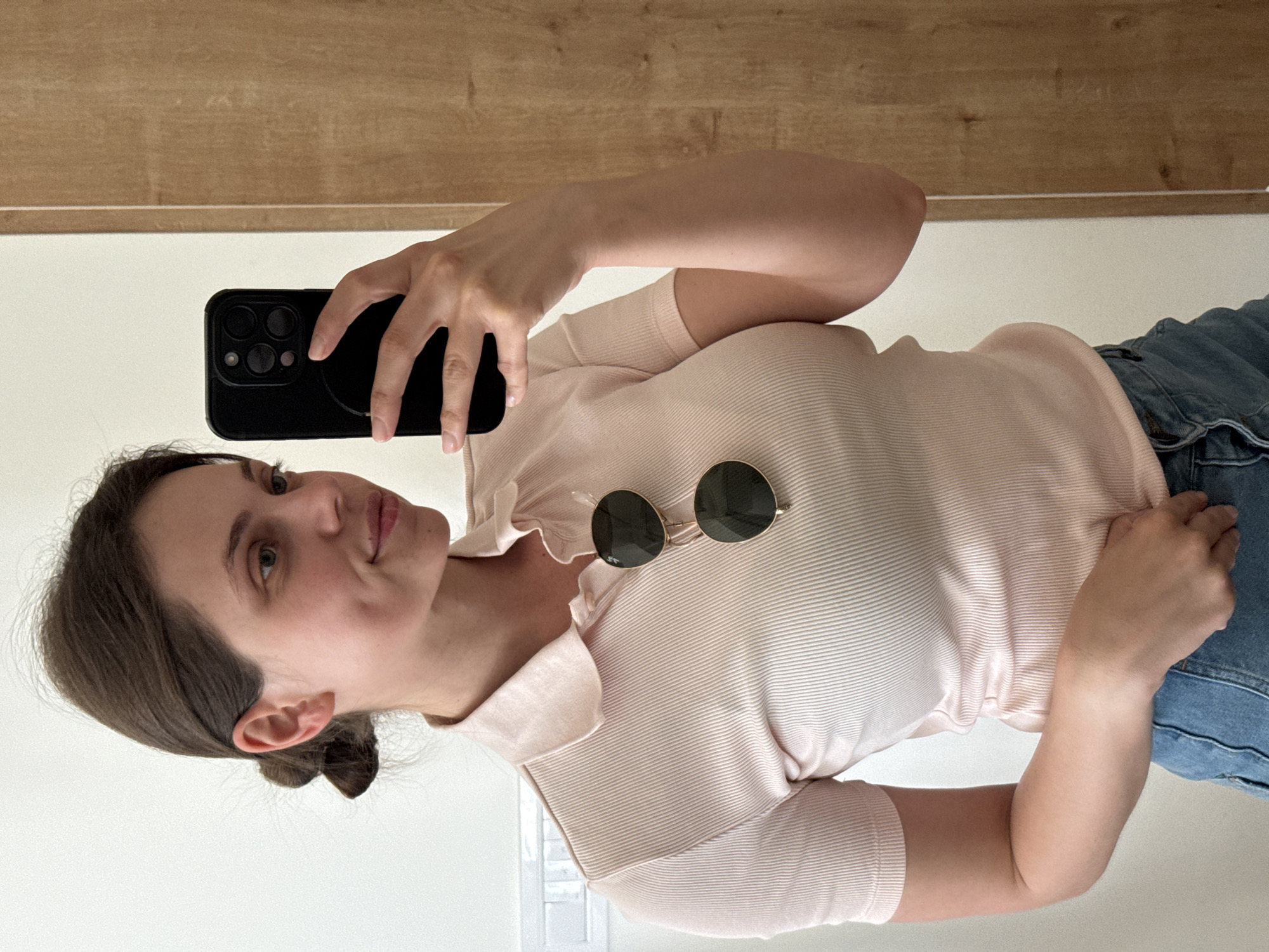 A woman is taking a selfie indoors, possibly in a mirror. She is holding a black smartphone with multiple camera lenses. She is wearing a light pink, ribbed shirt and jeans. A pair of sunglasses rests on her shirt. The background includes a wooden surface. The location, Belgrade, Serbia is not determinable from the image.
