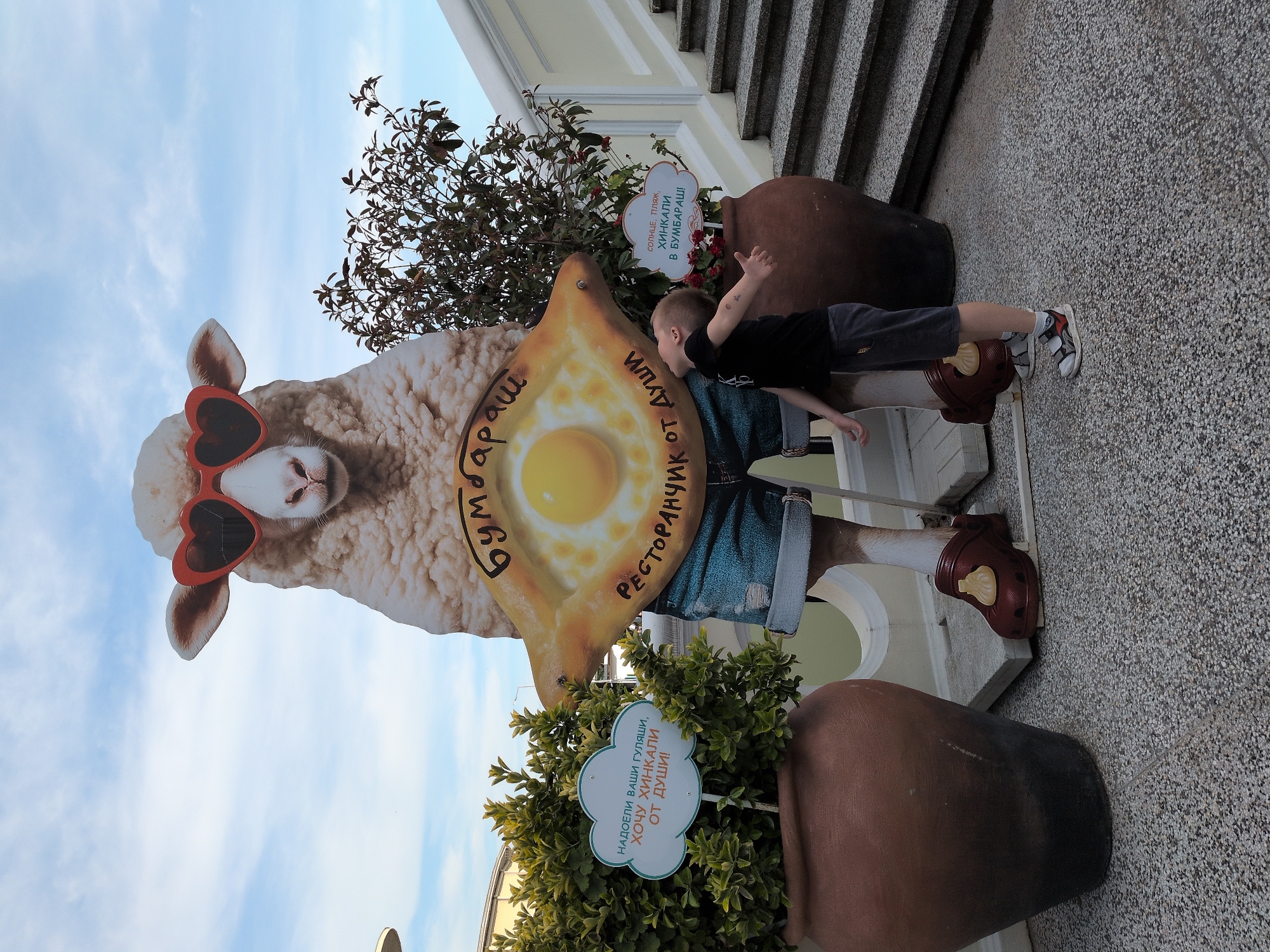 An outdoor scene in Sochi, Russia, features a large, whimsical cutout advertisement for a Georgian restaurant. The central subject is a life-sized depiction of a white sheep wearing red heart-shaped sunglasses, denim shorts, and brown clogs adorned with small khinkali dumplings. The sheep cutout holds a large, boat-shaped khachapuri with an egg in the center, displaying the text "БУМБАРАШ" (Bumbarash) and "РЕСТОРАНЧИК ОТ ДУШИ" (Restaurant from the heart).
To the right of the cutout, a young boy, approximately 4-7 years old, stands with his head tilted back, looking up at the sheep. He wears a black short-sleeved t-shirt, dark shorts, white socks, and grey/white sneakers. His arms are slightly extended outwards with palms facing upwards, indicating an inquisitive or appreciative posture towards the display.
Flanking the sheep cutout are two large, terracotta-colored planters. The left planter contains dense green foliage, while the right planter holds a shrub with darker green and reddish leaves, with some red flowers visible at its base. Each planter has a cloud-shaped sign with a light blue border. The sign in the left planter reads: "НАДОЕЛИ ВАШИ ГУЛЯШИ, ХОЧУ ХИНКАЛИ, ОТ ДУШИ!" (Tired of your goulash, I want khinkali, from the heart!). The sign in the right planter states: "СОЛНЦЕ, ПЛЯЖ, ХИНКАЛИ В БУМБАРАШ!" (Sun, beach, khinkali at Bumbarash!).
The ground is paved with light-colored, speckled aggregate. In the background on the right, a light-colored staircase with yellow railings ascends. To the left, a portion of a building and some urban infrastructure are visible. The sky above is light blue with scattered white clouds, suggesting clear daytime conditions.