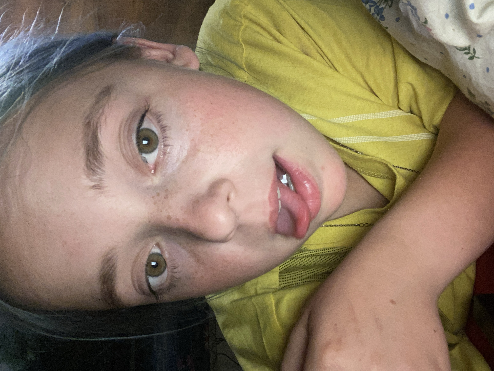 The image shows a close-up, rotated view of a young person lying in bed. The person has light skin with freckles across their nose and cheeks. Their eyes are wide and green, and their eyebrows are dark. The person's mouth is open, and their tongue is sticking out slightly. They are wearing a yellow t-shirt with thin white stripes. Part of a floral patterned sheet is visible behind them. The lighting suggests it is likely daytime, indoors. The overall impression is playful and candid. There is no visible text in the image.