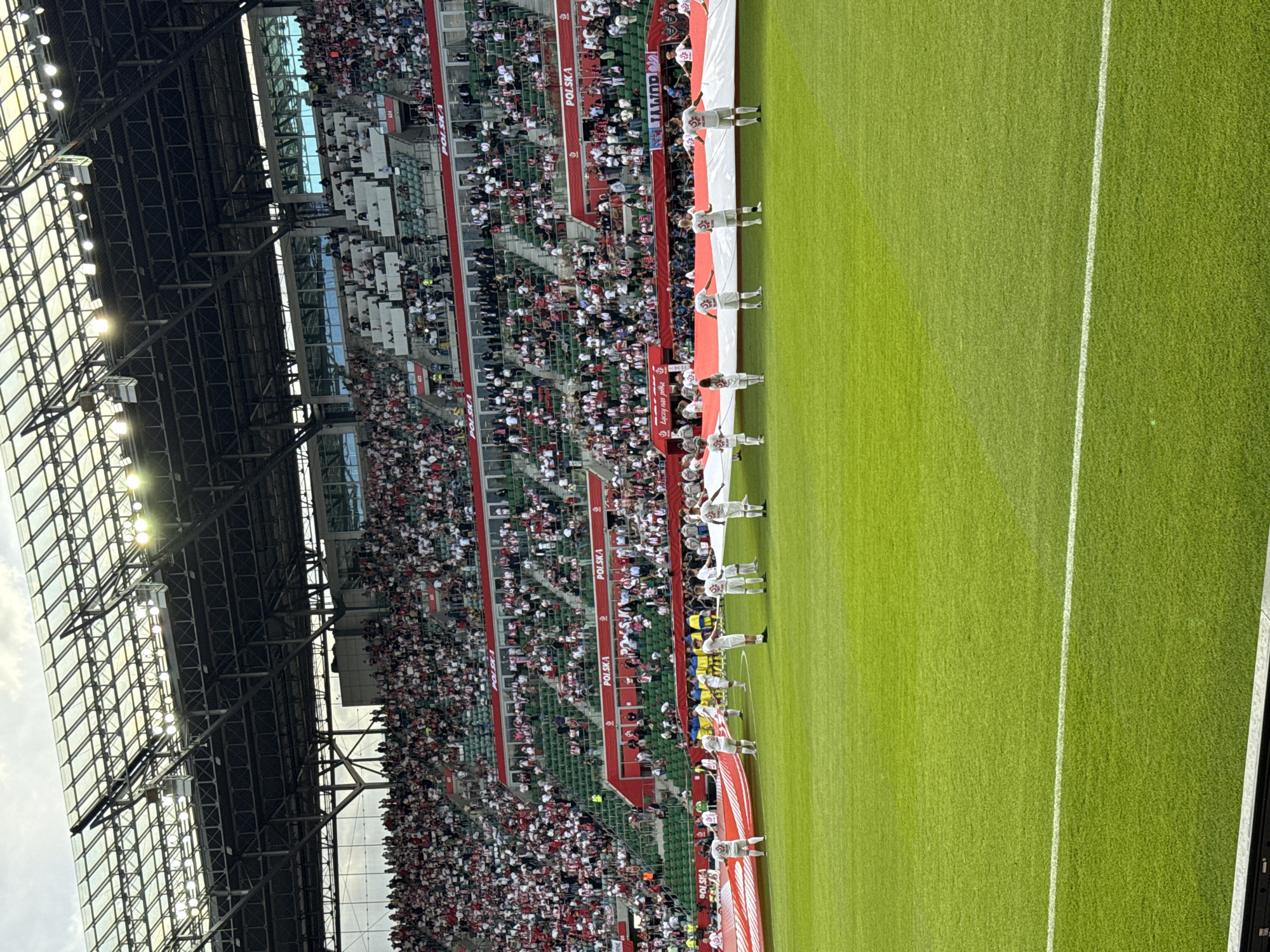The image captures a soccer stadium filled with spectators. The scene is likely set in Poland, as indicated by the "POLSKA" signage on the stadium walls. The stadium is crowded with fans, predominantly dressed in red and white, the national colors of Poland. Along the edge of the field, a group of people stand holding a large banner or flag. The stadium has a modern architectural design, with a visible metal framework and a partial glass roof. The soccer field is green with white lines marking the boundaries. The lighting suggests that the event is taking place during daylight hours, possibly late afternoon or early evening.