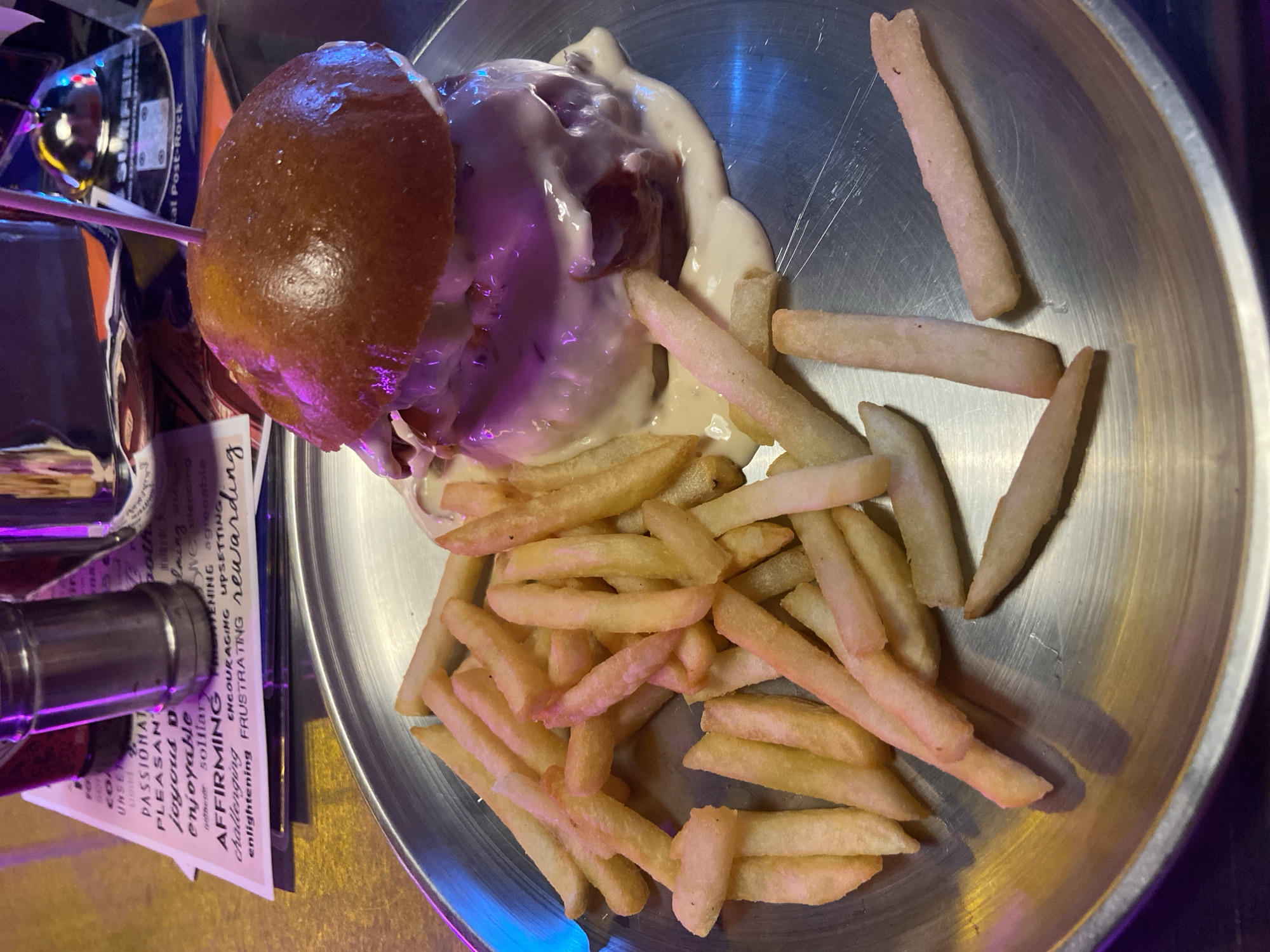 The image shows a close-up of a hamburger and french fries served on a metal plate. The hamburger is topped with a golden-brown bun and covered in a white sauce, with hints of red showing through. A pink toothpick is stuck in the top bun. The french fries are golden yellow and appear to be thick-cut. The plate is placed on a surface with some blurred text in the background that appears to be on a menu or placemat. The lighting suggests an indoor environment, possibly a restaurant, with purple and blue accents from ambient lighting. No people are visible in the image. There is no information in the image to suggest its location in Bac Nha Trang Ward, Vietnam.