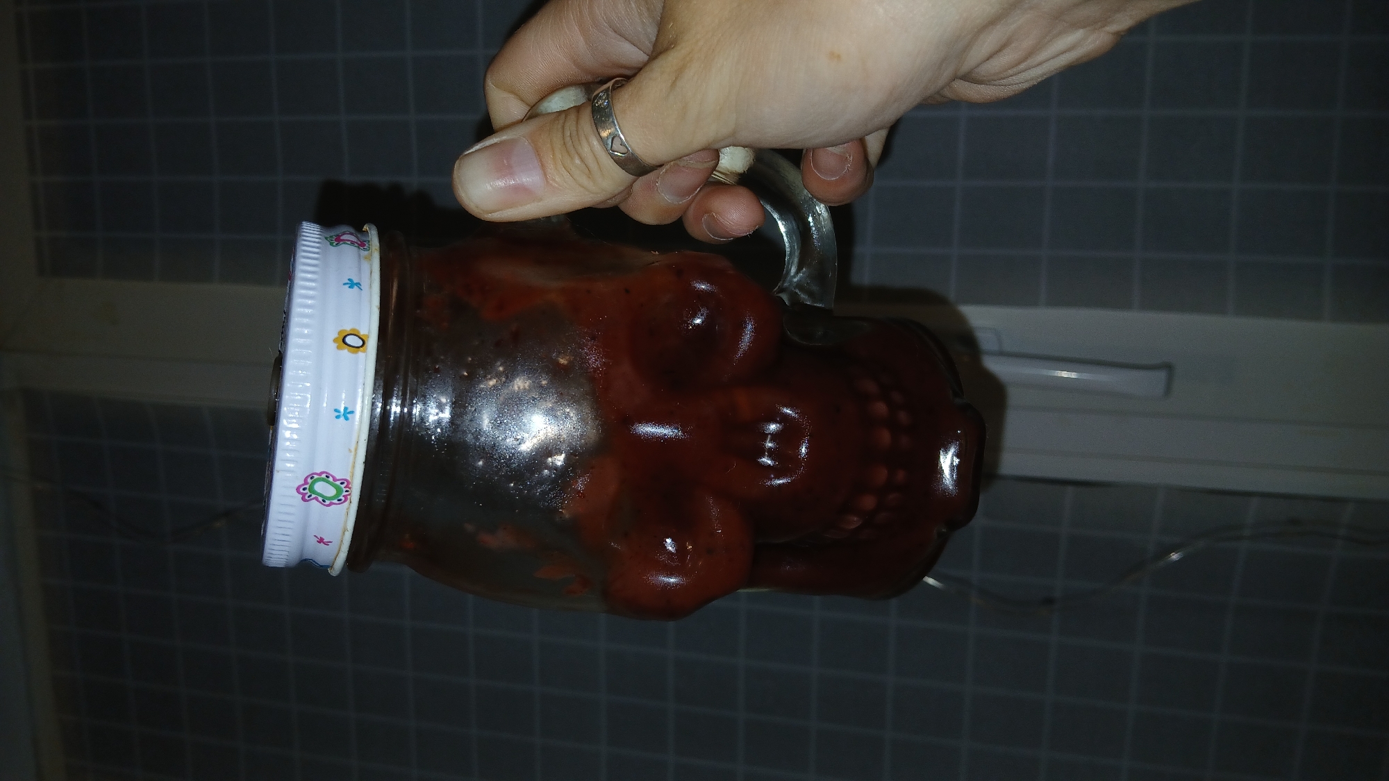 A hand holds a skull-shaped glass mug filled with a dark red substance. The mug has a handle and a white lid with floral decorations. The hand has a silver ring with a heart on the ring finger. The background is a grid surface. The location is unknown.