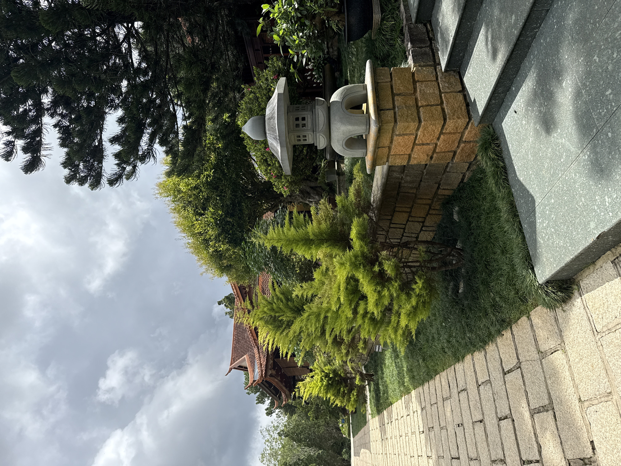 The image depicts a serene courtyard with a stone pathway leading past a miniature Zen garden. In the foreground, a stone lantern sits atop a decorative brick pedestal, flanked by lush green shrubs and meticulously manicured grass. The pathway, paved with light-colored flagstones, winds towards a traditional Vietnamese building with a terracotta-tiled roof, visible in the background. The building's architecture, characterized by its ornate eaves and wooden structures, suggests a temple or a cultural heritage site. Towering pine trees, with their dark green foliage, contrast with the vibrant green of the garden, and the sky above is overcast with dramatic, heavy clouds, hinting at an impending rain. This tranquil scene is characteristic of the many spiritual and cultural sites found in Da Lat, Vietnam, a city renowned for its cool climate and natural beauty.