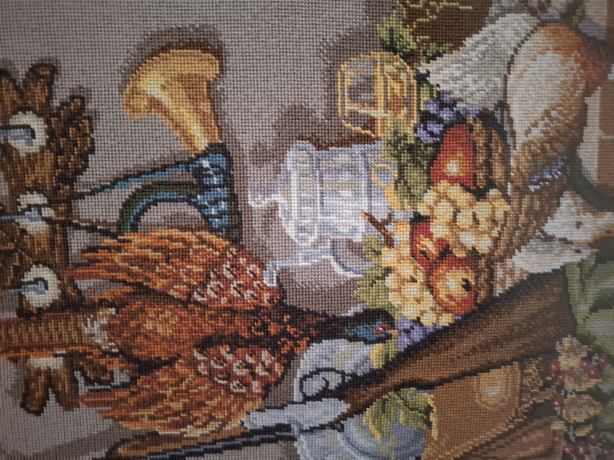 This is a close-up, eye-level, medium shot of a cross-stitch tapestry. The tapestry depicts a still life composition, possibly a hunting or harvest theme, with various objects arranged together.
Dominating the upper left is a large, ornate bird with intricate feather detail in shades of orange, brown, and white. To its right, a golden horn-like object juts out. Below these, a light blue object with black detailing hangs.
In the central area, a white, detailed pitcher or jug with a lid and handle is prominent. To the right of the pitcher, a yellow, rectangular object with dark lines, possibly representing a cart or wagon, is visible.
The foreground is filled with a basket overflowing with fruit, including apples in shades of red and orange, and bunches of grapes in purple and yellow. To the left of the basket, a portion of a rifle or gun is visible, with its stock and barrel. Behind the gun, a brown bag or pouch with a flap closure is present. A white object, possibly a bird or small animal, is positioned in the lower right corner, partially obscured by the fruit basket. Green foliage and other smaller decorative elements are scattered throughout the scene. The background is a neutral, woven texture in shades of gray and beige, characteristic of cross-stitch fabric.
There are no people visible in the image. The objects are static and arranged in a decorative manner. The environment is an indoor still-life as depicted in a textile art piece. There is no information in the image to associate it with Nordhausen, Germany.