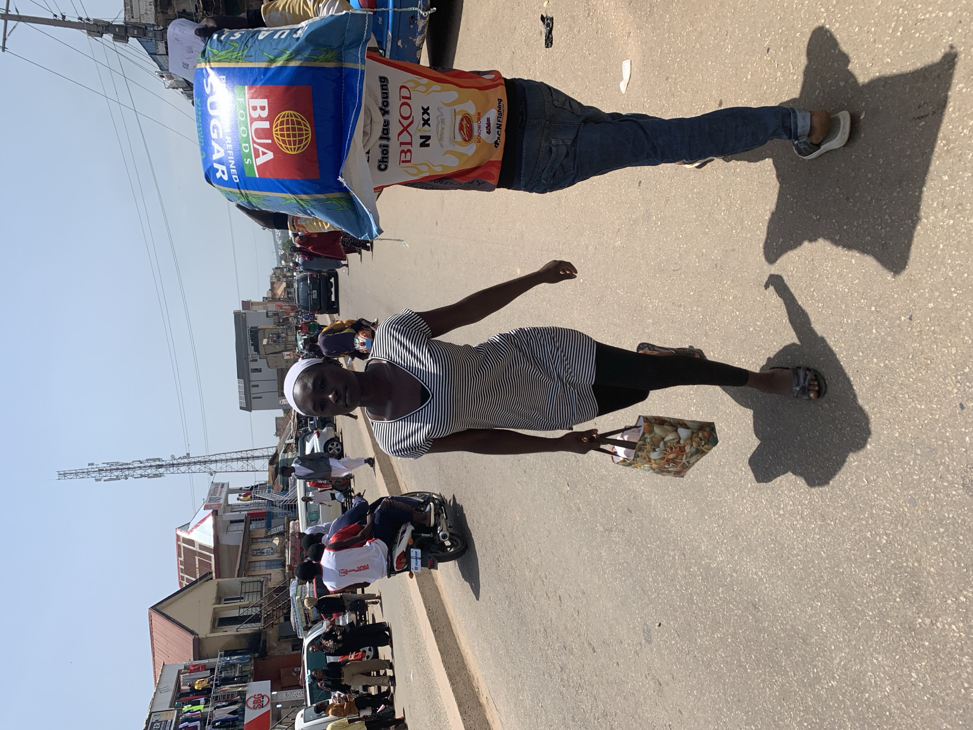 In Jos, Nigeria, a busy street scene unfolds under a bright sky. A young woman, wearing a striped dress and a white headscarf, walks towards the camera, carrying a patterned shopping bag in her right hand. Behind her, a man carries a large, blue and red bag of BUA refined sugar on his shoulders. He is wearing a t-shirt with the text "Choi Jae Young BIXOD N XX" and blue jeans. In the background, a motorcycle with three passengers is visible, along with other pedestrians and vehicles on the asphalt road. Buildings line the street, displaying various businesses and wares, including clothing hanging outside. A telecommunications tower stands tall in the distance. The overall atmosphere suggests a typical day in a bustling Nigerian marketplace.