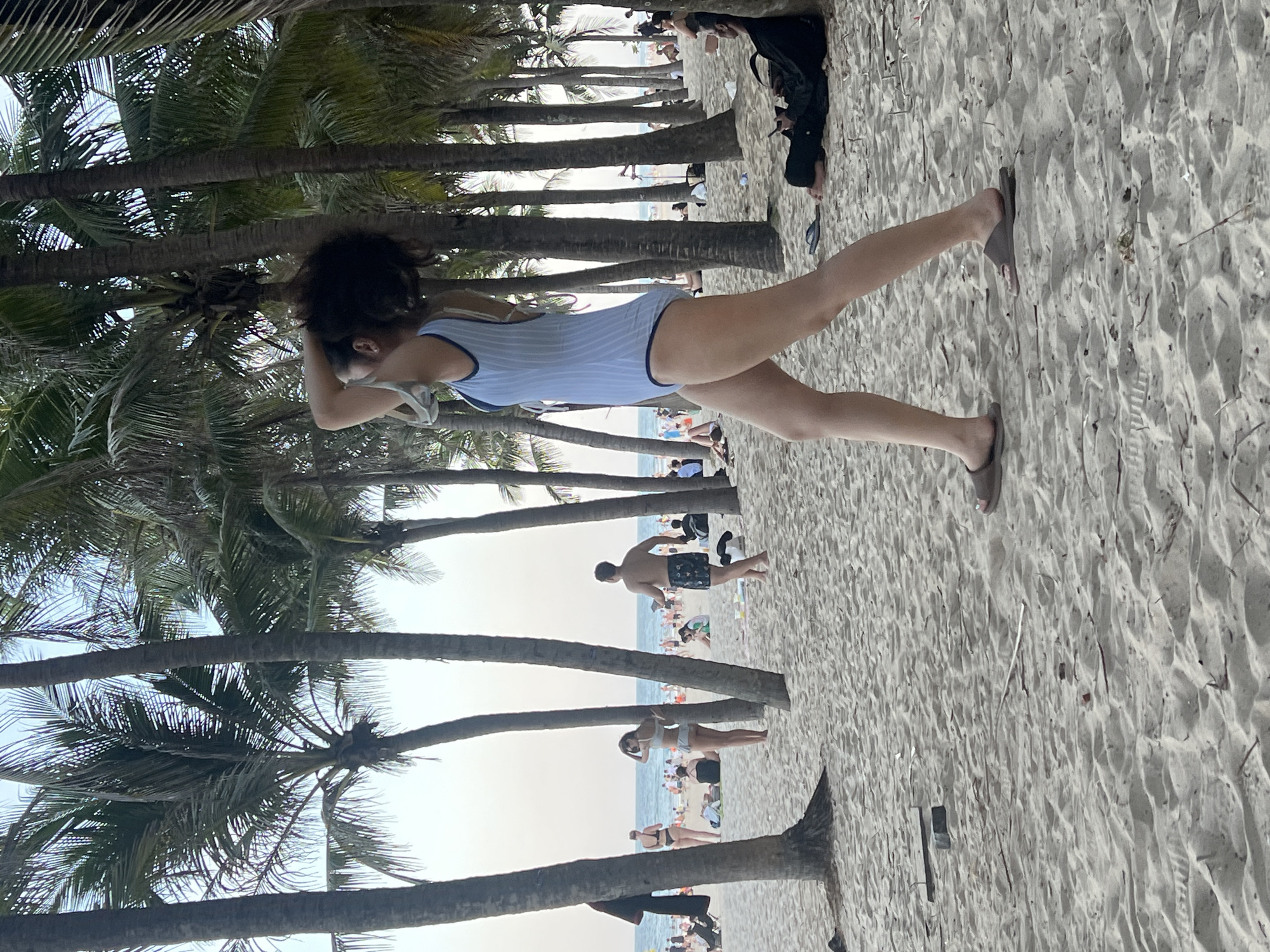 A woman in a light blue one-piece swimsuit walks on a sandy beach lined with palm trees. She has dark, curly hair and is seen from the side, looking to her left. She is wearing brown sandals and has her right arm raised, touching her hair. In the background, other beachgoers are visible, some sitting on towels and others walking. The ocean is in the distance under a hazy sky. The scene appears to be at My Khe Beach in Đà Nẵng, Vietnam, known for its wide sandy shores and palm trees.
