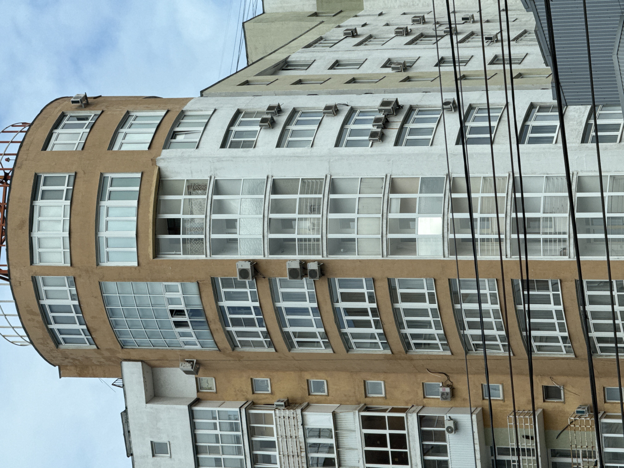 The image shows a close-up, low-angle view of a modern, multi-story apartment building with a curved facade in Chelyabinsk, Russia. The building is a mix of light tan and white concrete, with numerous large windows and balconies. Several air conditioning units are mounted on the exterior walls. Overhead power lines are visible in the foreground, obscuring parts of the building. The sky is overcast. The architecture suggests a relatively recent construction, typical of urban residential developments in Russia.