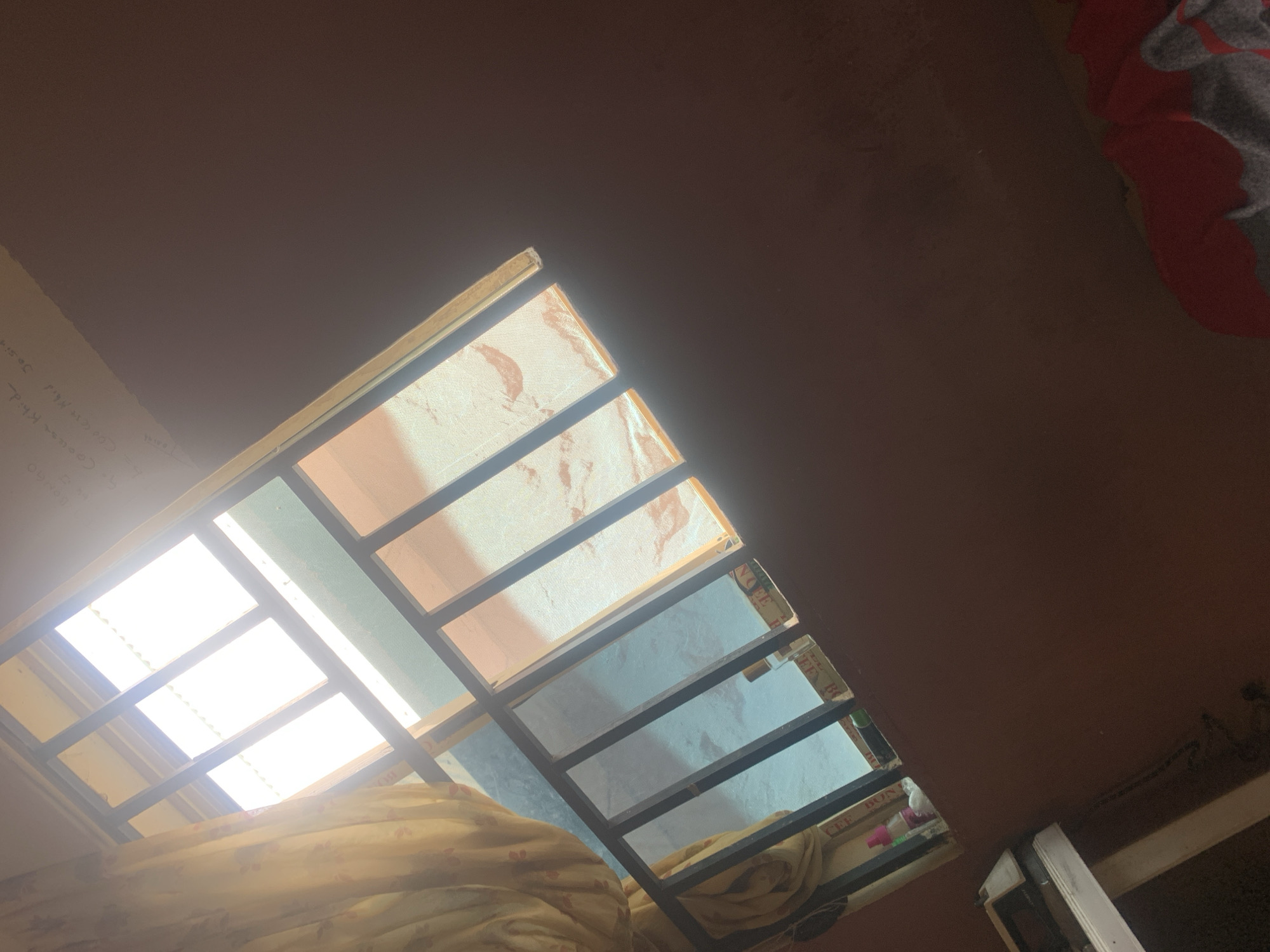 The image shows a room with a window covered by a yellow curtain and bars. Sunlight streams through the window, illuminating the room. There are some writings on the wall above the window. The room appears to be in a residential setting in Jauro Yinu, Nigeria. The walls are painted a dark reddish-brown. In the foreground, a piece of furniture, possibly a table or desk, is visible, with some items on it. A red and gray blanket is partially visible in the bottom right corner.